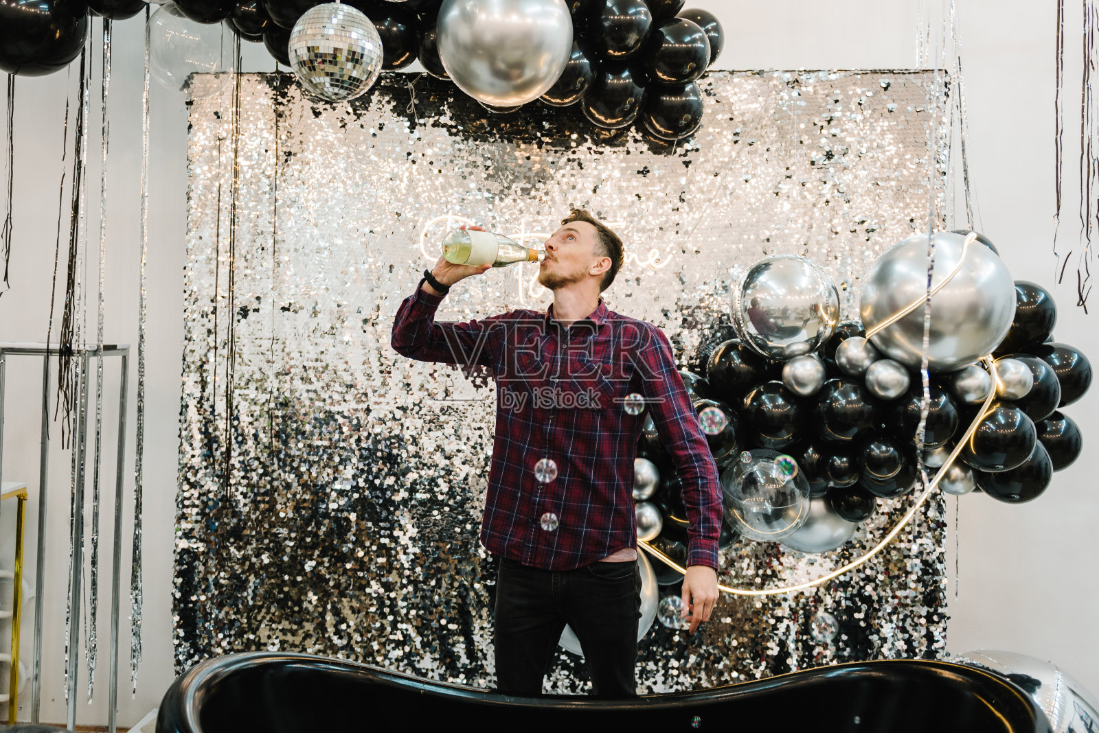 快乐男子喝香槟庆祝2023年。祝贺生日的地方。拱门或照相亭装饰黑色银色气球。墙，带浴室的摄影区。派对时间。照片摄影图片