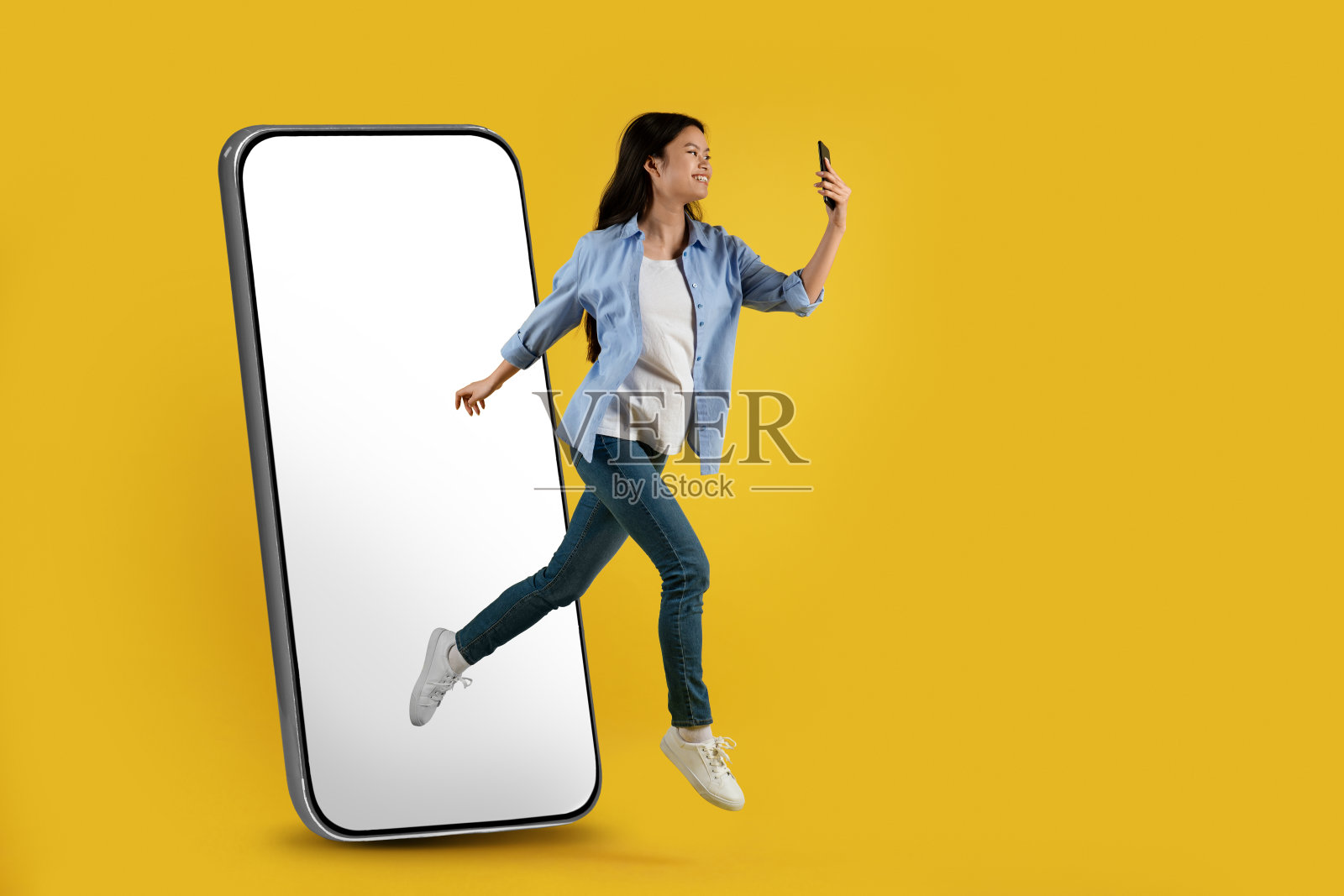 满意的年轻亚洲女士从巨大的空白屏幕的手机上跳下来，冻结在空气中，看着智能手机照片摄影图片