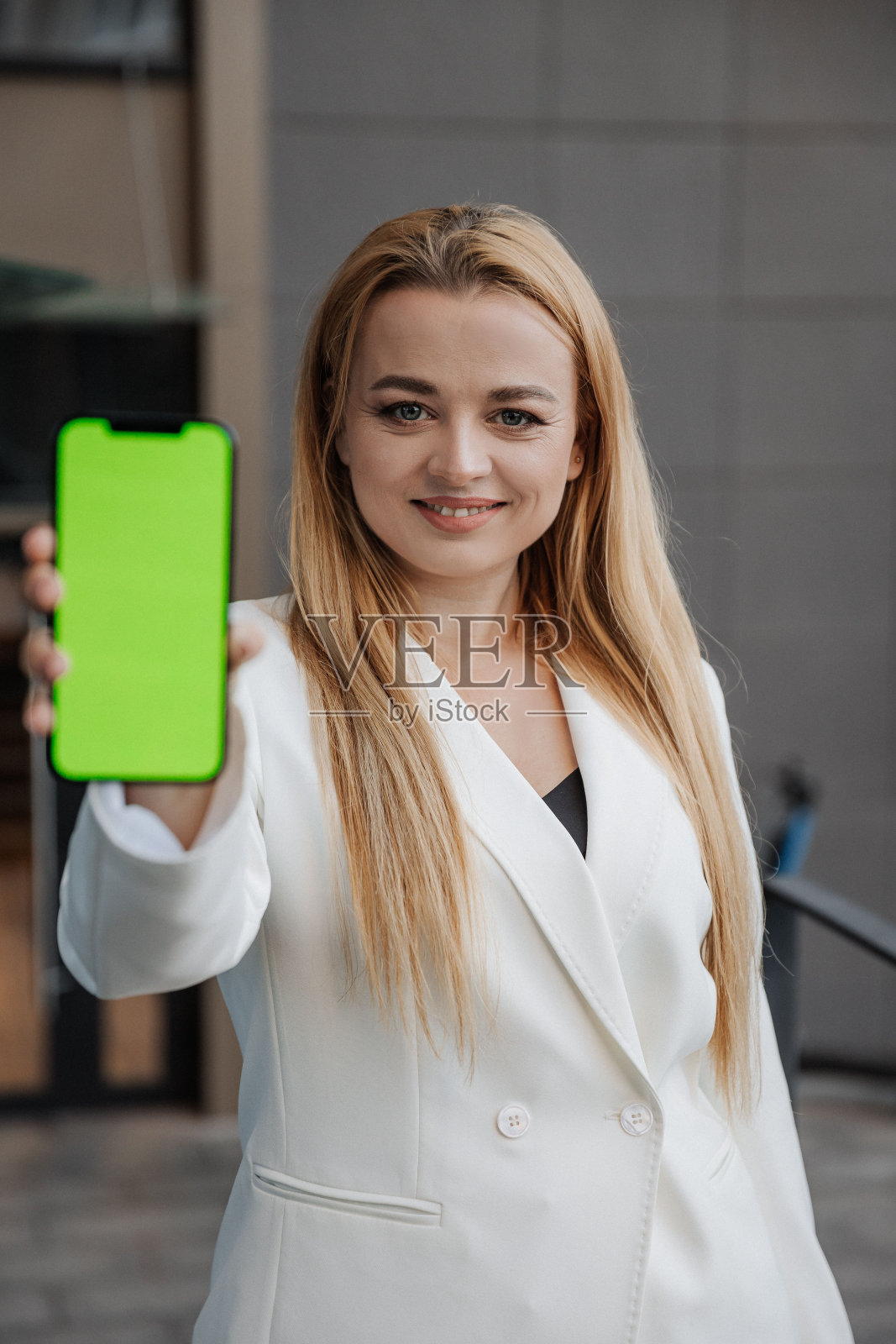 金发微笑的女人在超大的白色夹克显示手机屏幕上的城市背景铬照片摄影图片