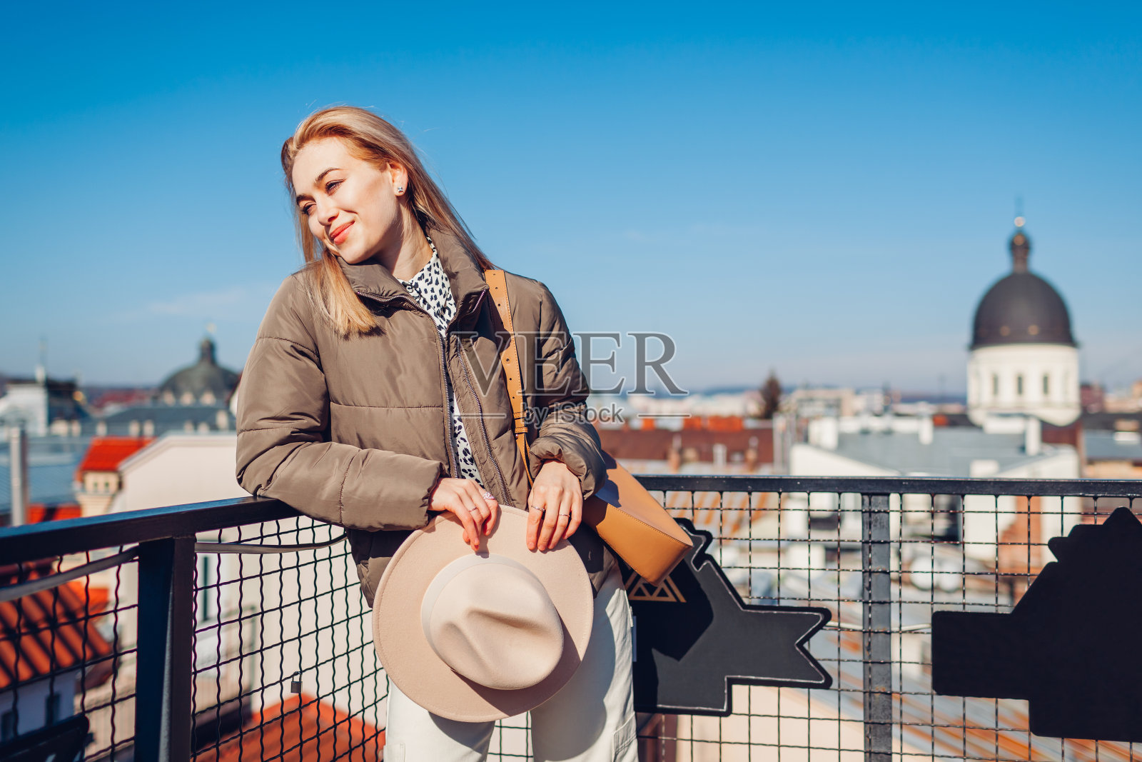 时尚的年轻女子从欧洲城市的阳台上拿着帽子欣赏风景。春季时装照片摄影图片