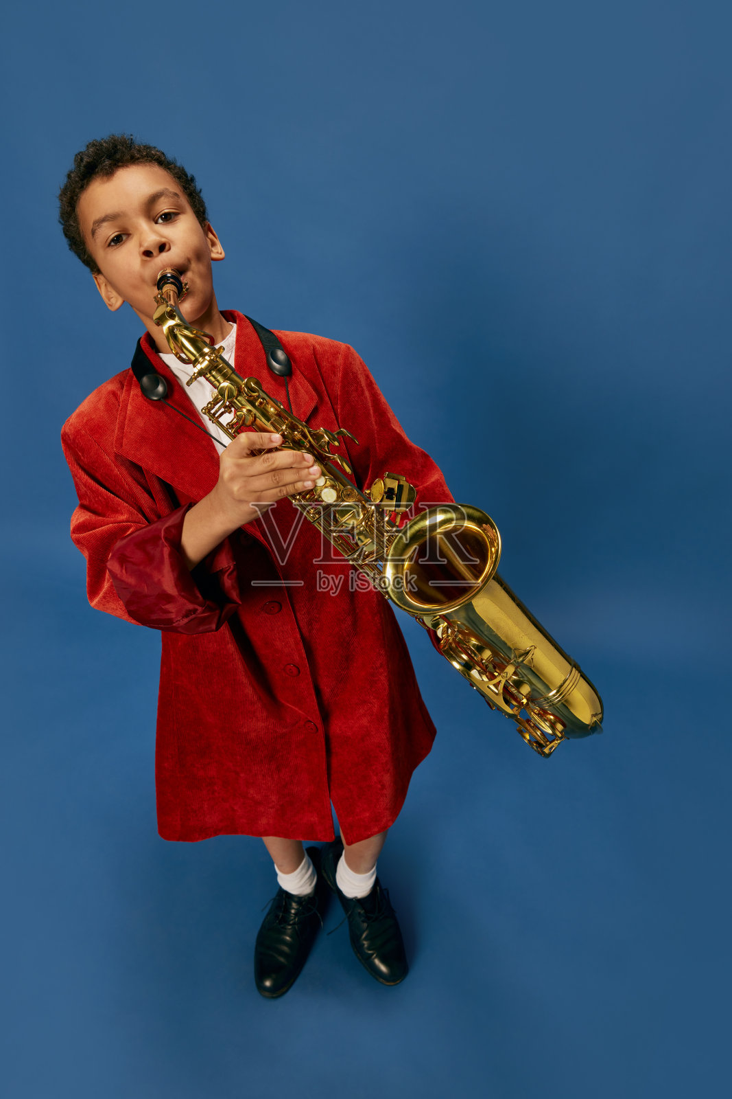 可爱的非洲小男孩穿着巨大的男人夹克和鞋子，就像爵士男人在蓝色背景上演奏萨克斯。时尚，艺术，音乐和创意风格照片摄影图片