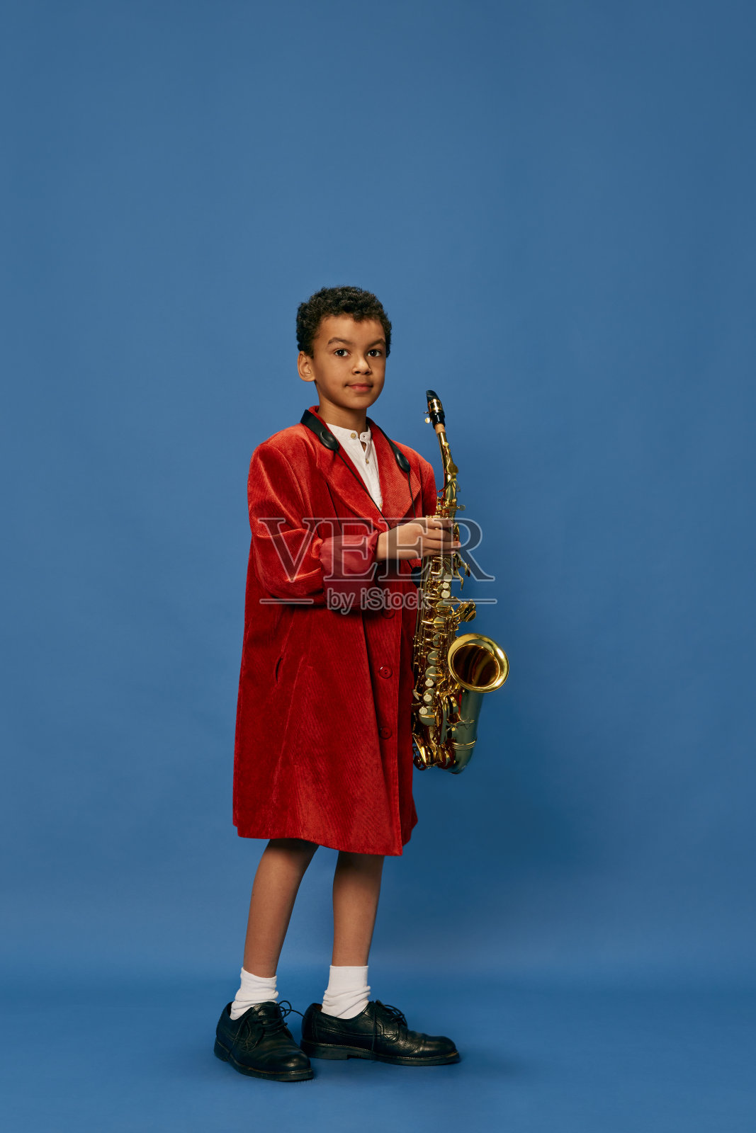 可爱的小非洲男孩穿着巨大的男子夹克和鞋子的肖像，就像爵士男人在蓝色背景上演奏萨克斯。时尚，艺术，音乐和创意风格照片摄影图片