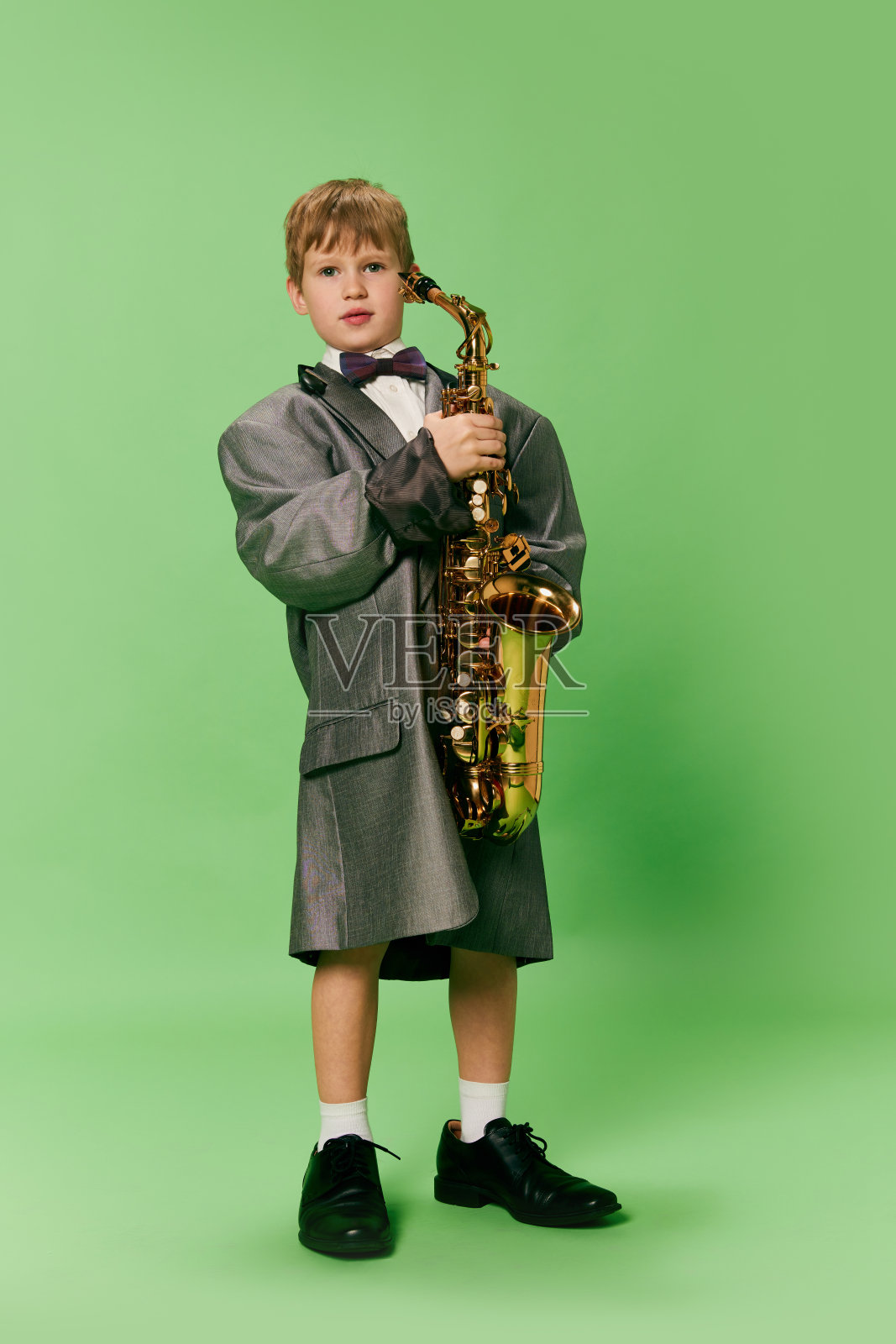 时尚的小学龄男孩在巨大的超大复古风格的衣服和大的男性鞋摆姿势与尤克里里吉他孤立在绿色背景。照片摄影图片