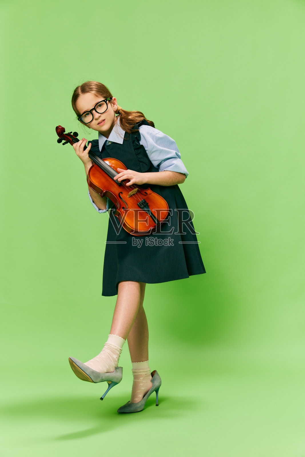 可爱的小女孩，才华横溢的音乐家穿着巨大的母亲的高跟鞋拉小提琴，有乐趣孤立在绿色的背景。教育、音乐、经典艺术、新视野、趣味理念照片摄影图片