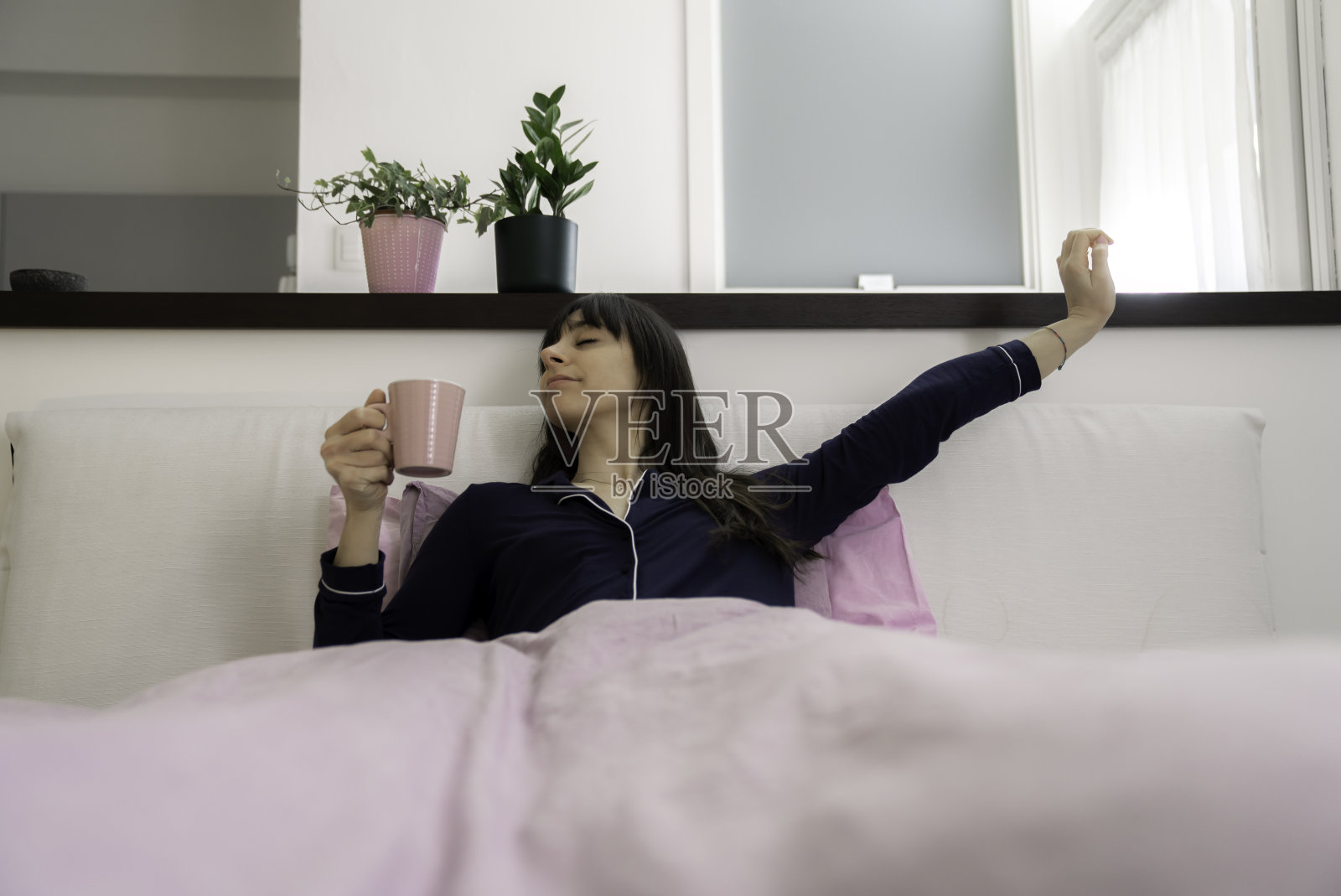 漂亮的年轻女人躺在床上喝咖啡或茶，用笔记本电脑在家远程办公，早上醒来时伸展身体照片摄影图片