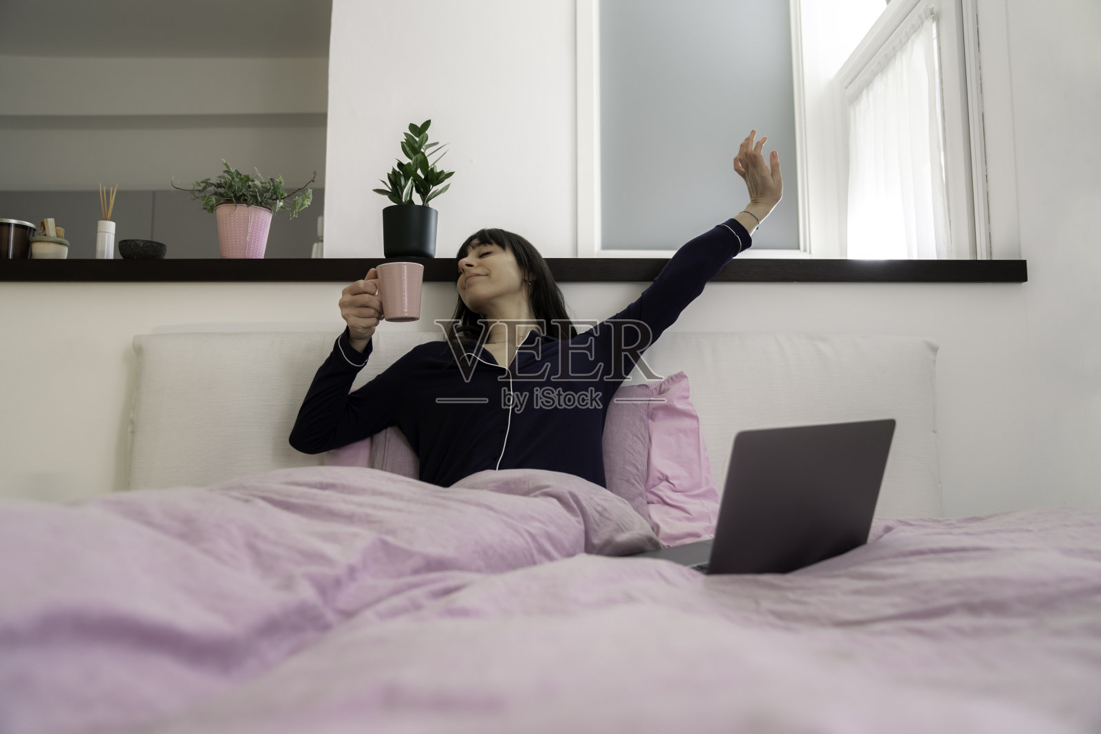 漂亮的年轻女人躺在床上，穿着睡衣喝着咖啡或茶，拿着笔记本电脑在家远程办公，早上醒来时伸展身体照片摄影图片