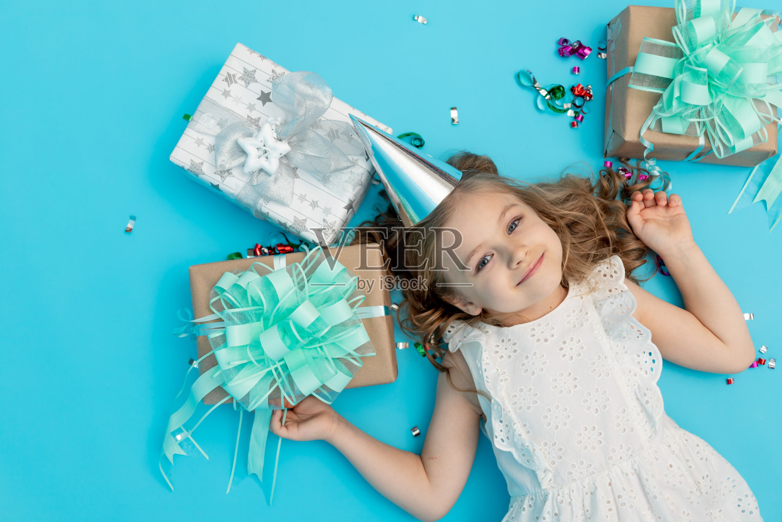 一个可爱快乐的小女孩穿着蓝色背景的纯棉白色连衣裙，戴着节日帽子，上面有礼物和五彩纸屑，她正在庆祝她的生日，这是一个发短信的地方照片摄影图片