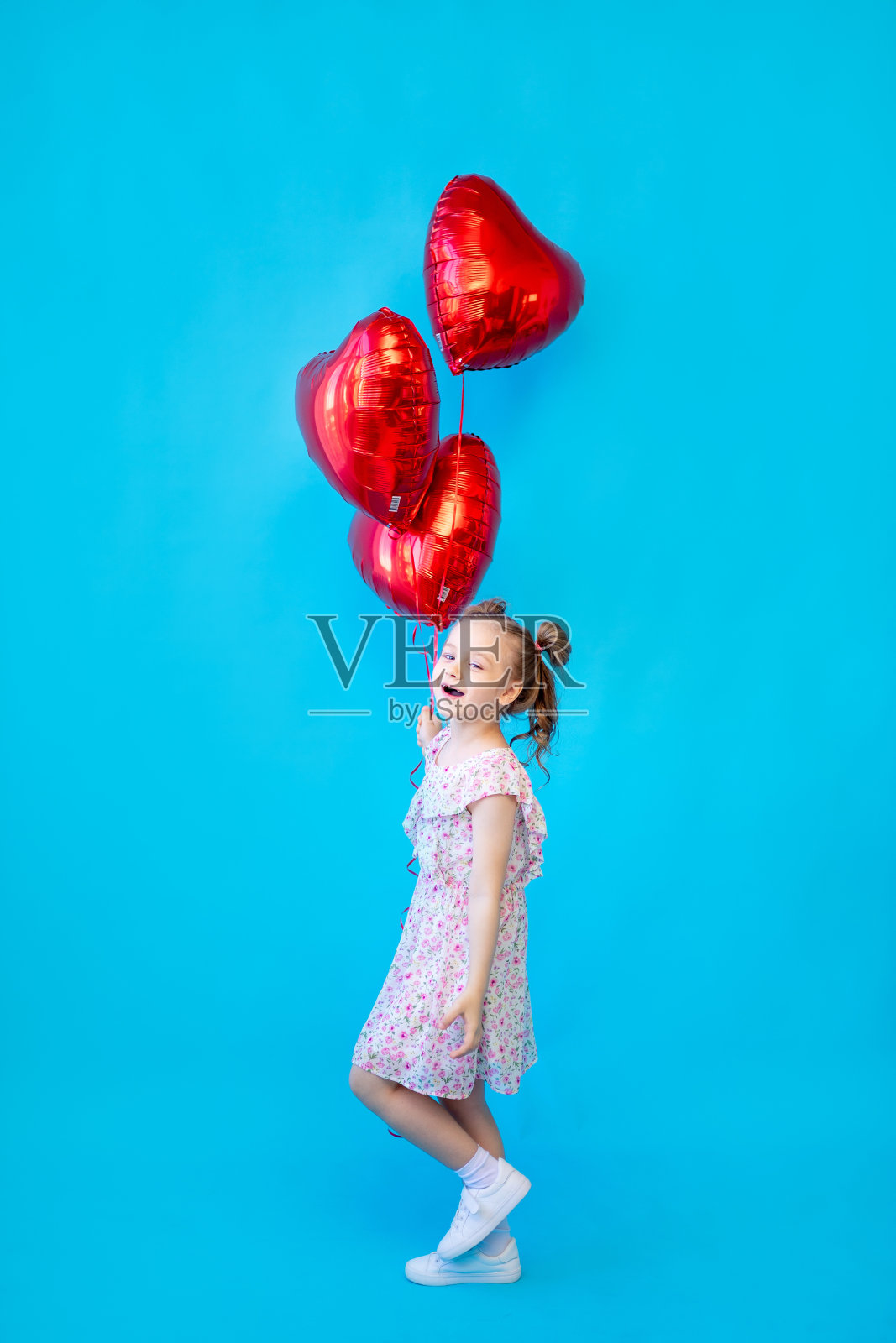 一个小女孩在蓝色的孤立工作室背景上，红色的气球在一个心形的地方玩得很开心。用于复制文本的空间。情人节概念照片摄影图片
