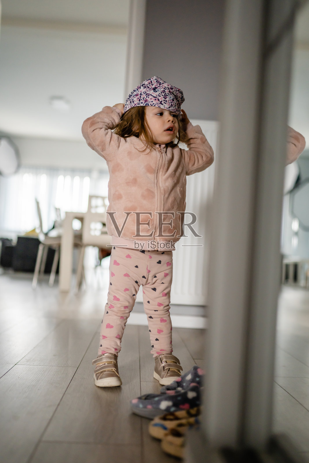 一个蹒跚学步的女孩站在家里穿衣，试着把帽子戴在头上，独立的孩子和早期儿童发展的概念照片摄影图片