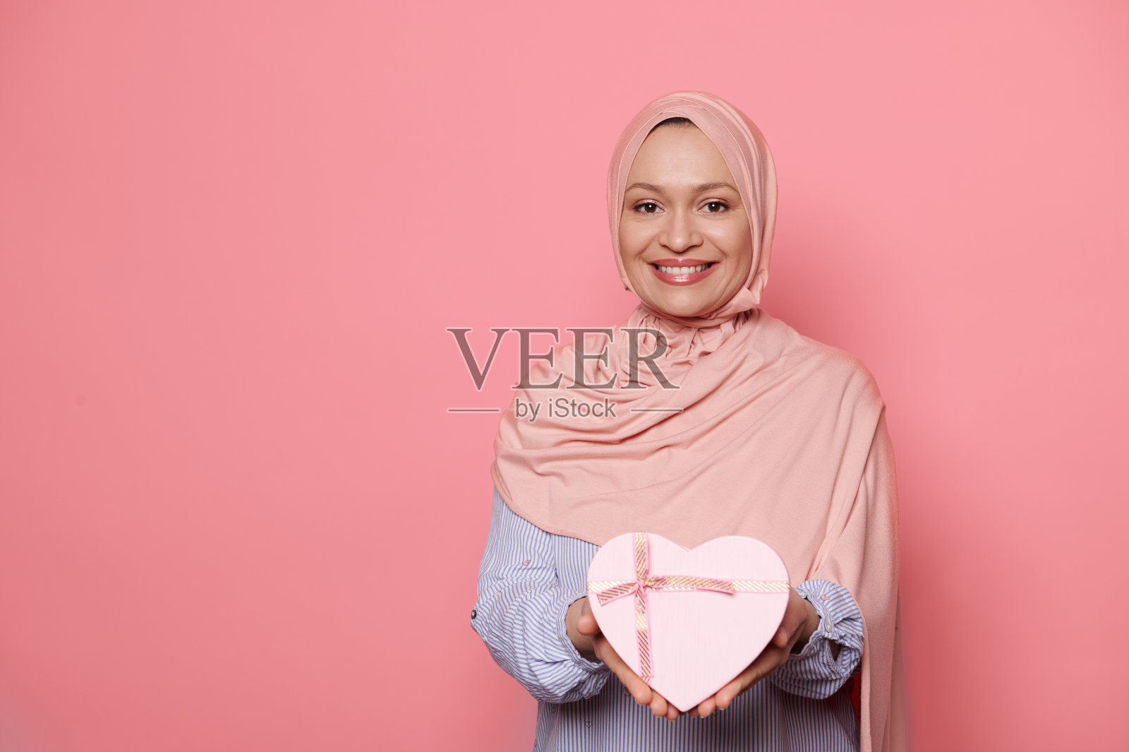中年穆斯林妇女微笑着，在镜头前拿出一个可爱的心形粉色礼盒，孤立在粉红色的背景上照片摄影图片