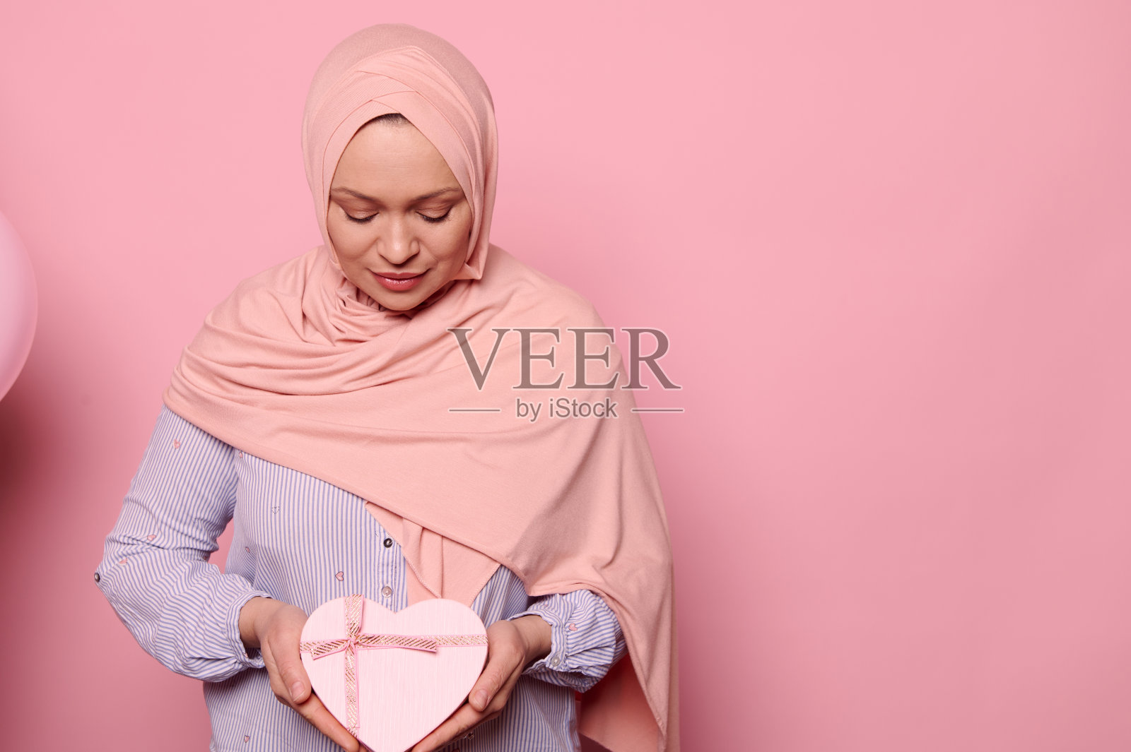 华丽的穆斯林妇女在头巾和蓝色衬衫，拿着心形的粉色礼盒里面的惊喜，孤立在粉红色照片摄影图片