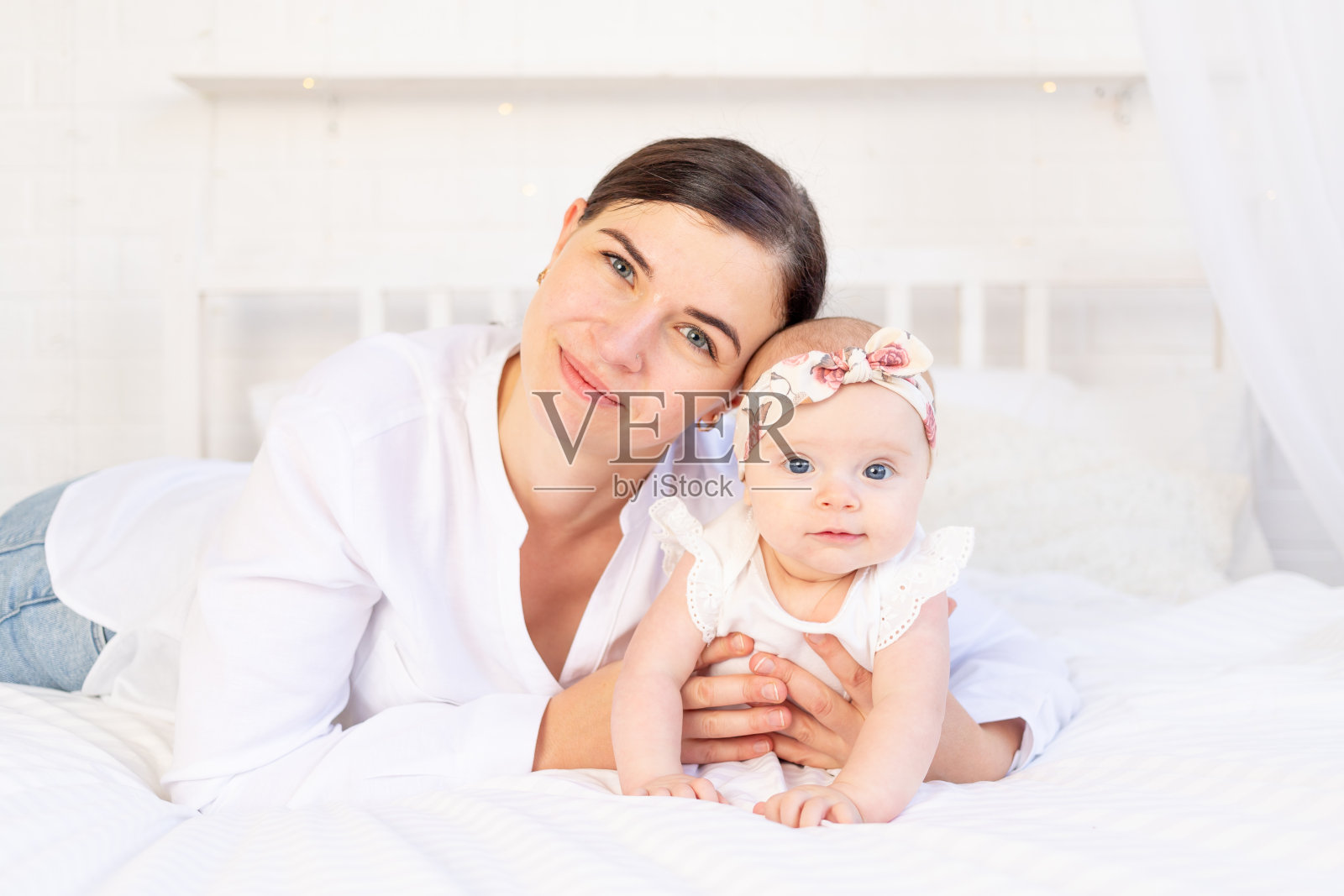 妈妈和女婴抱在一起躺在家里的白色棉床上，母爱和关怀照片摄影图片
