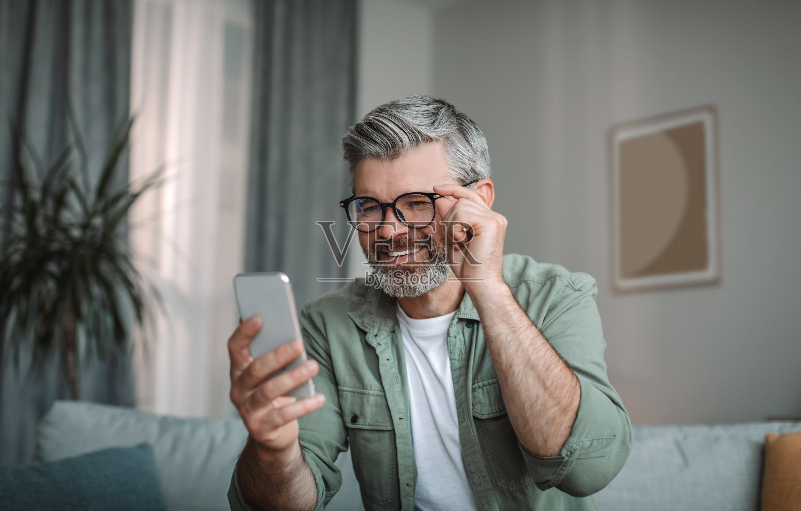 高兴的戴眼镜留着胡子的白人老人男性看着智能手机，在房间内部得到了好消息，复制空间照片摄影图片
