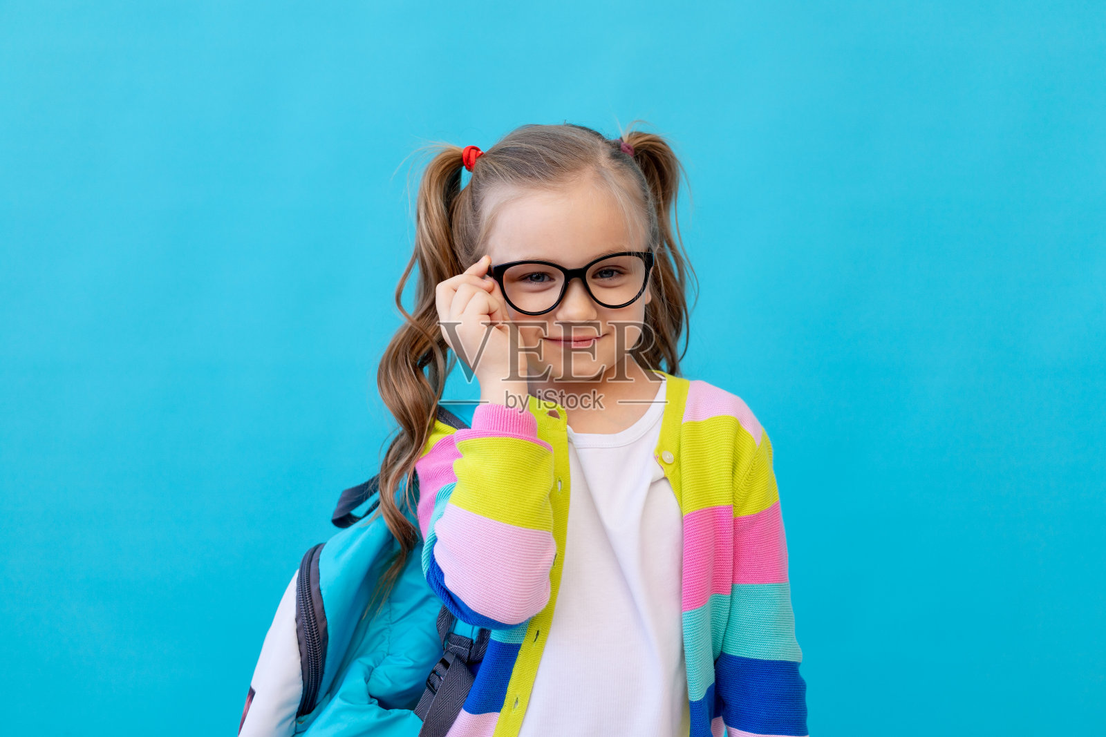 一个可爱的小女孩戴着眼镜，穿着条纹夹克，带着笔记本和背包，像老师一样调整她的眼镜。教育理念。照相馆，蓝色背景，文字的地方照片摄影图片
