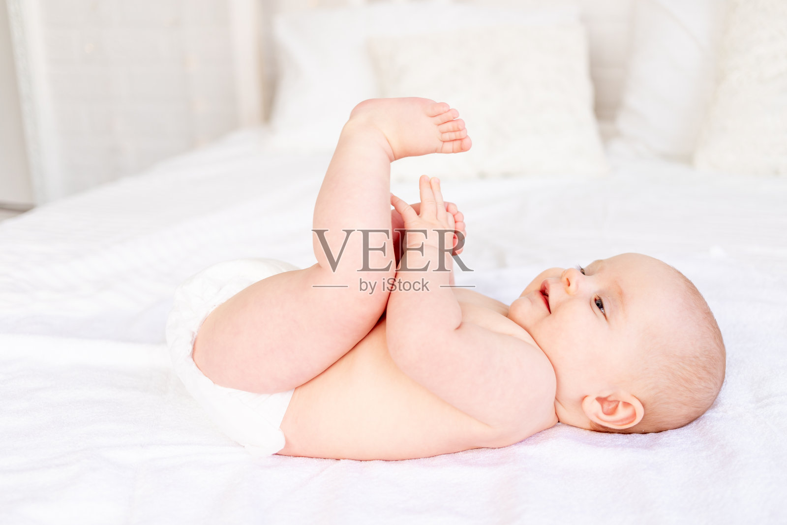 快乐而欢笑的宝宝在尿布里玩脚，在白色棉床上的婴儿床上，卫生和新生儿护理照片摄影图片