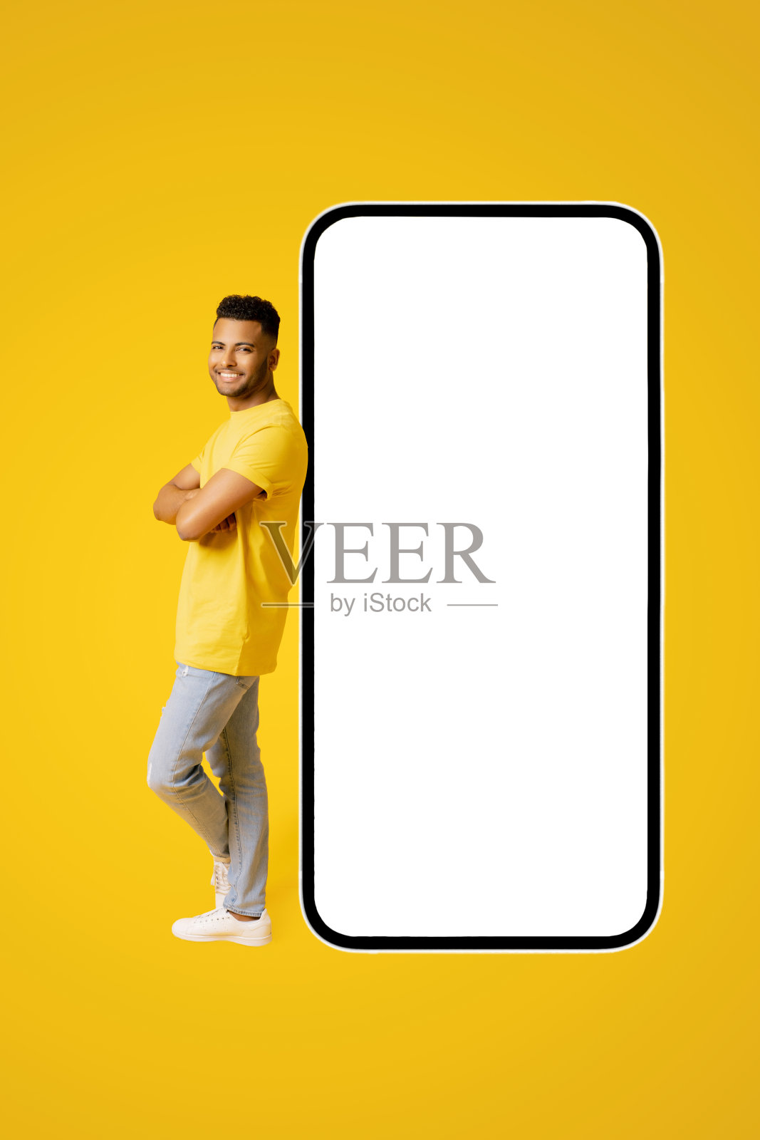 开朗自信的印度年轻人靠在巨大的智能手机上，黄色背景上空屏幕隔离，阿拉伯人双臂交叉，展示和广告移动应用程序，全长照片摄影图片