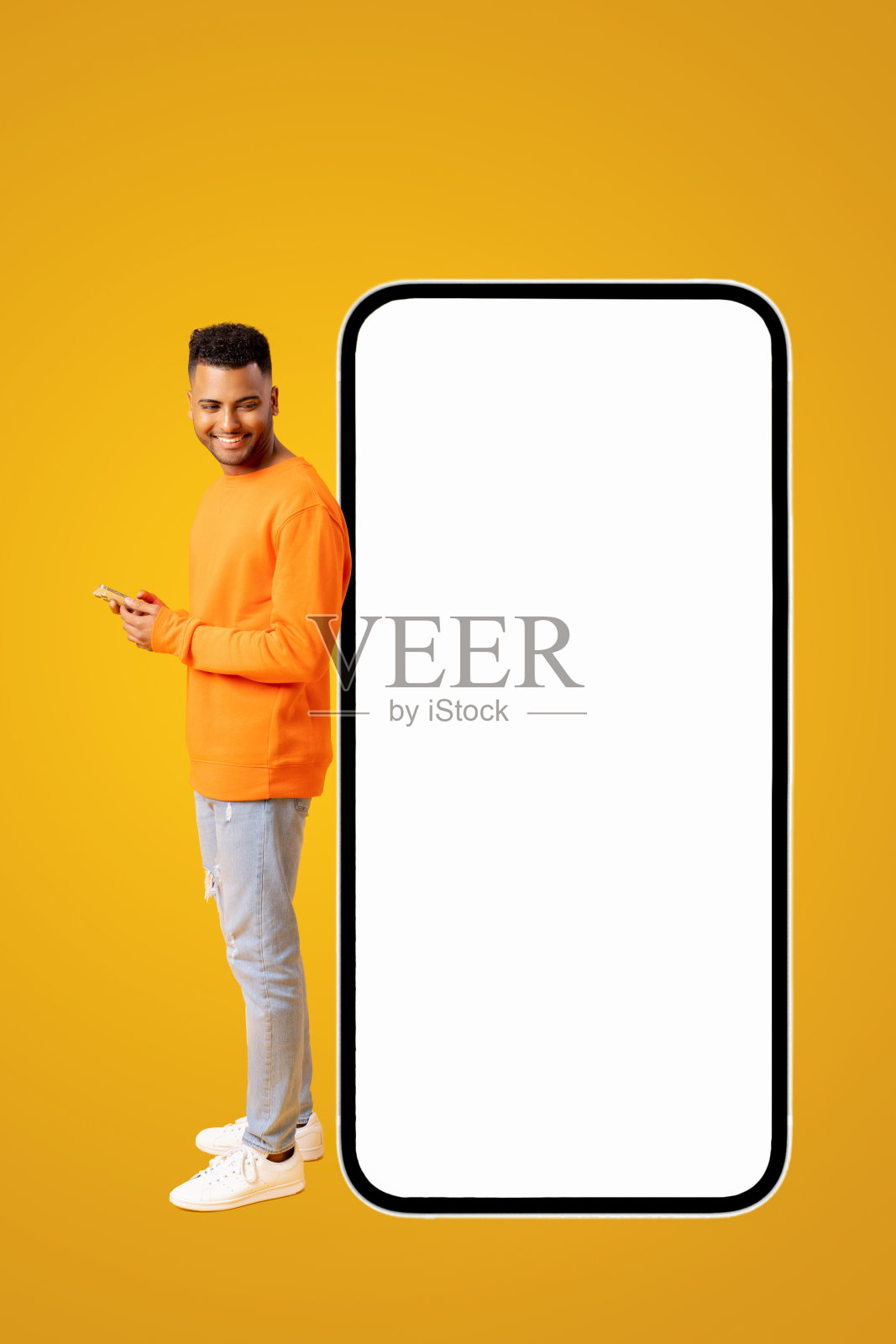 穿着休闲装的微笑迷人的印度男人站在巨大的智能手机旁边，看着屏幕，使用手机，潮人在宣传新的应用程序或网站，模型照片摄影图片