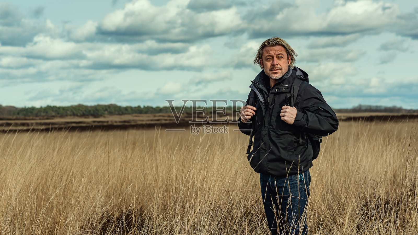 在多云的天空下，一个金色头发，黑色夹克，背包的男人在荒地上。照片摄影图片