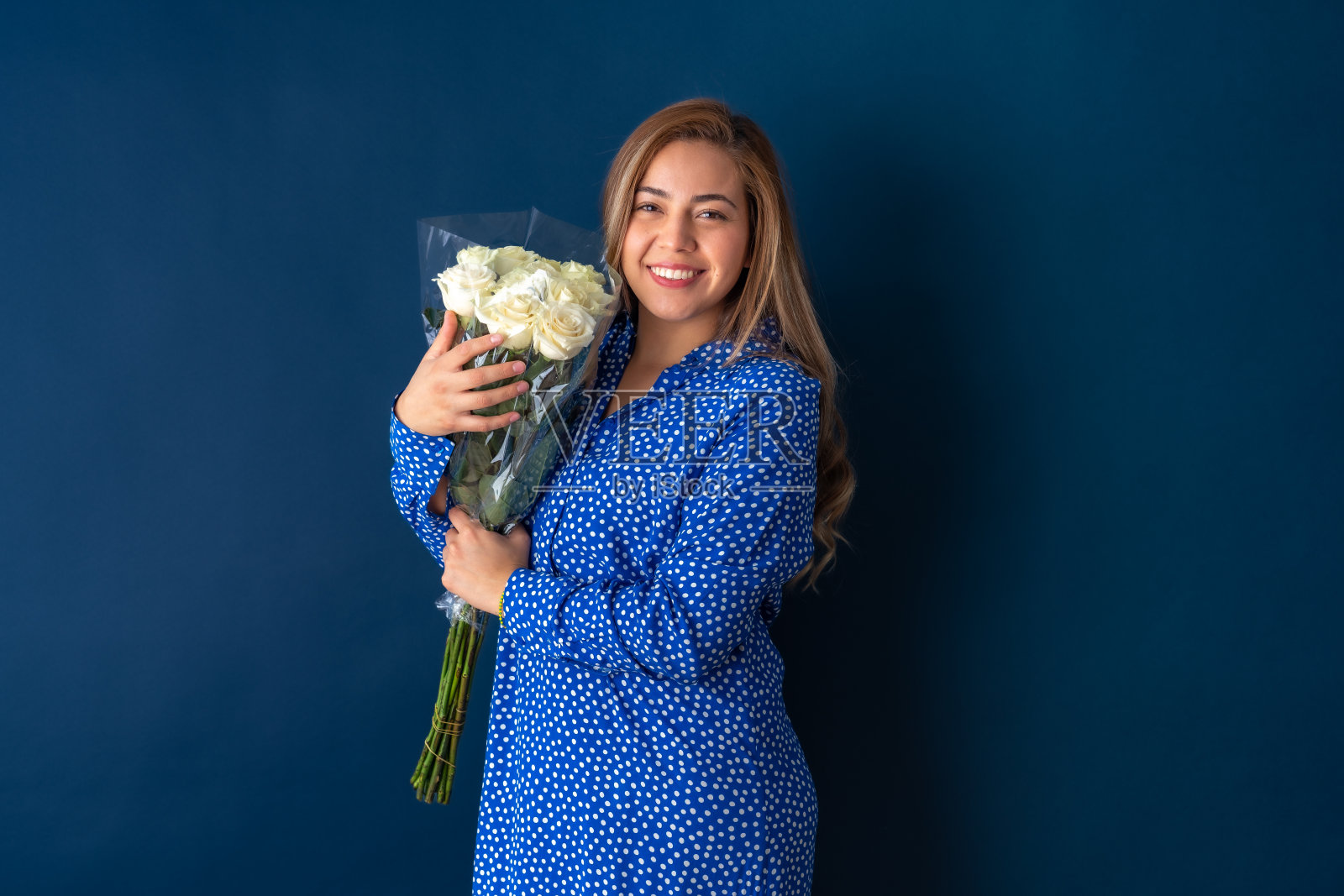 美丽迷人的年轻女子在爱情中快乐地摆姿势，手持一束白玫瑰，穿着蓝色的点裙。在蓝色背景上隔离。照片摄影图片