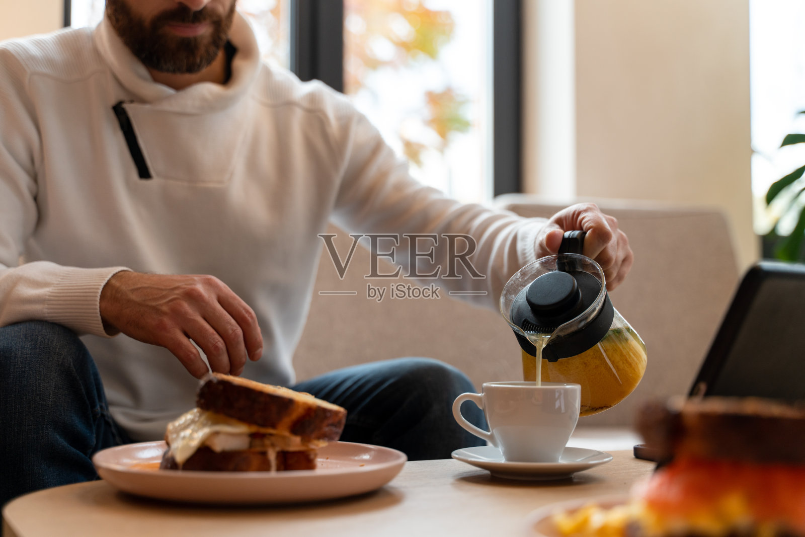 英俊的年轻人坐在桌子上的平板电脑和工作。他正在往杯子里倒咖啡照片摄影图片