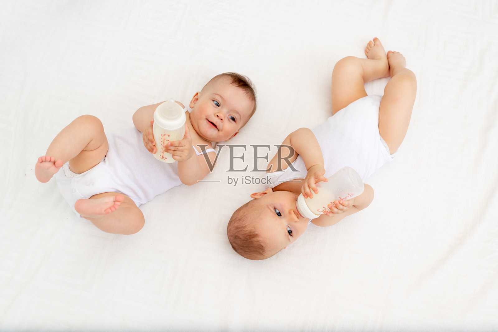 在儿童房的白色床上，两个婴儿躺在婴儿床上，从一个瓶子里吮吸牛奶照片摄影图片
