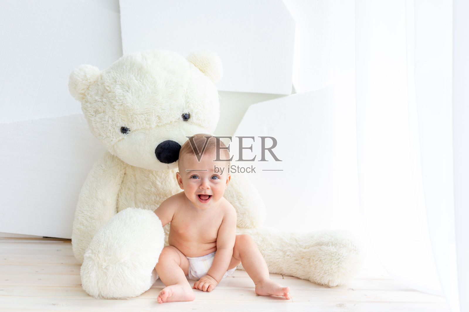 一个6个月大的小女孩穿着尿布和一只柔软的大熊坐在明亮的公寓里照片摄影图片