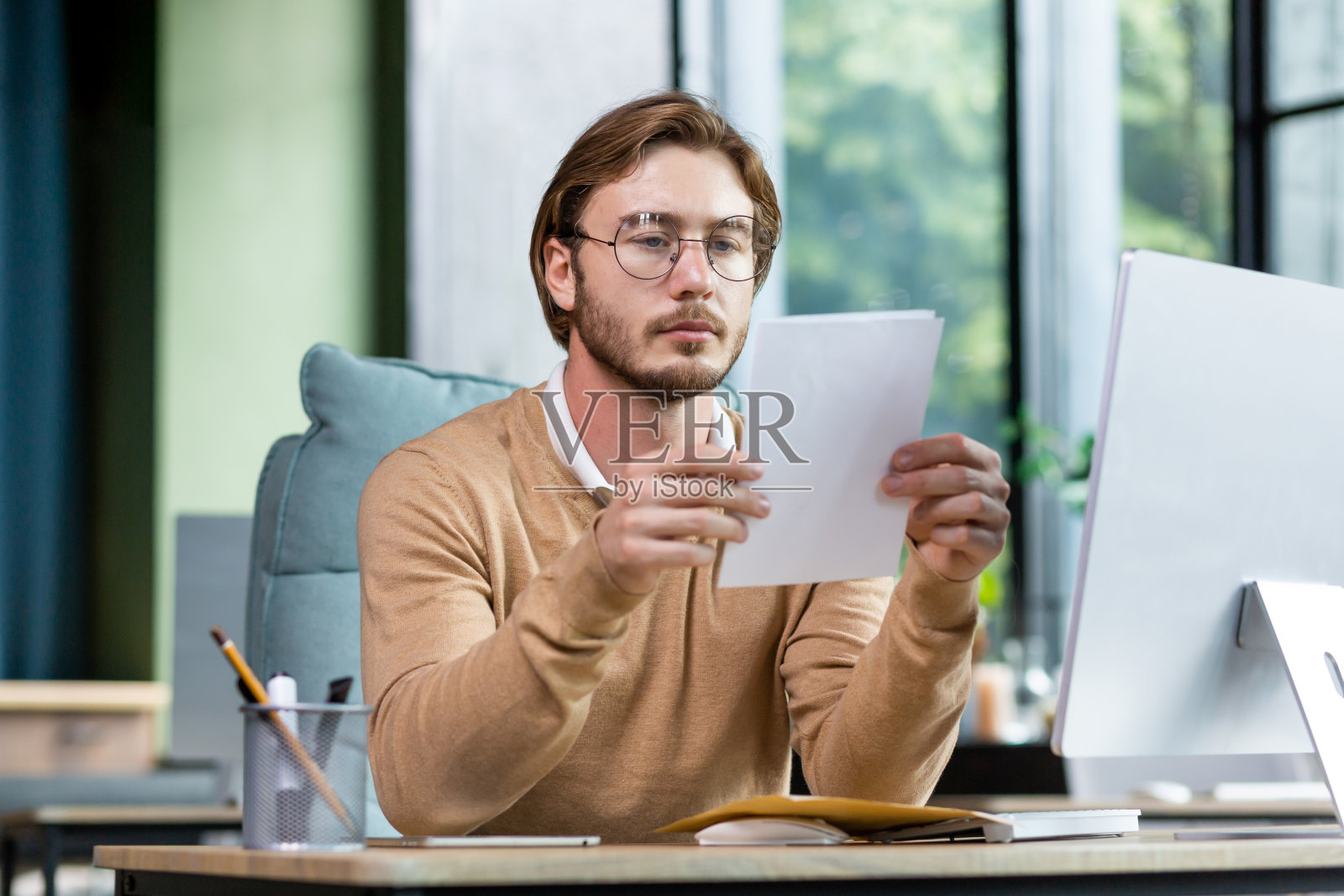 一个严肃的年轻人商人，自由职业者，程序员坐在办公室的桌子前阅读文件，信件照片摄影图片