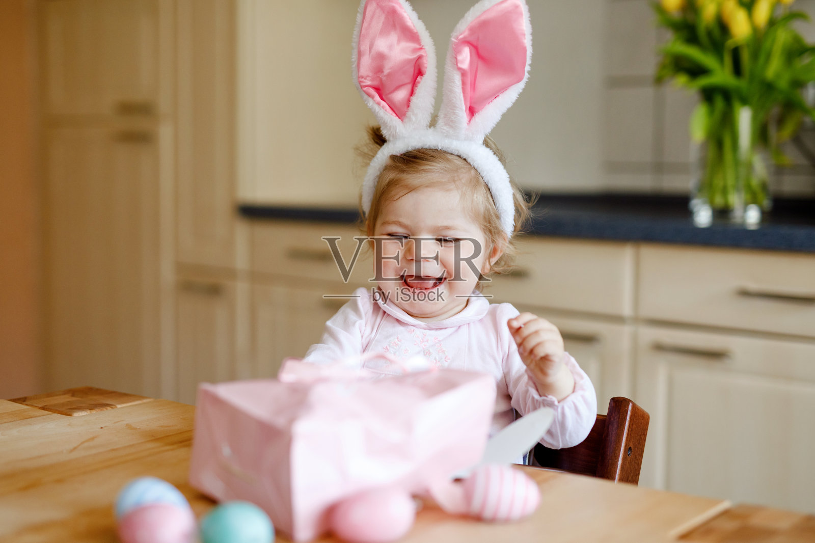 可爱的小蹒跚学步的女孩戴着复活节兔子耳朵玩彩色粉彩蛋。快乐的宝宝孩子拆礼物。可爱的健康微笑的孩子在粉红色的衣服享受家庭假期照片摄影图片