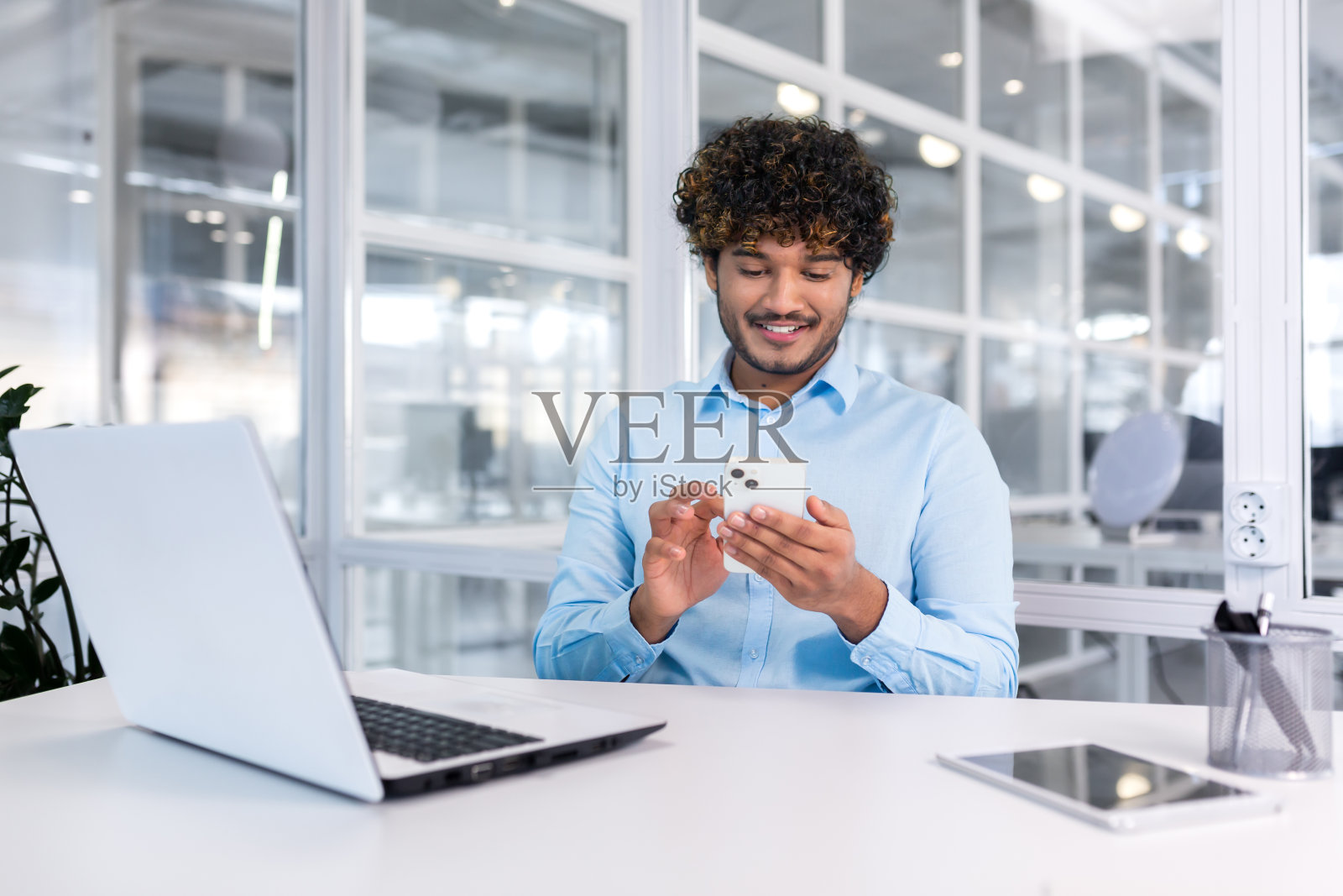 年轻的印度程序员在现代办公室里坐在工作场所，男人用电话，测试新软件的在线应用程序，穿着衬衫的商人企业家微笑着，为取得的成就而高兴照片摄影图片