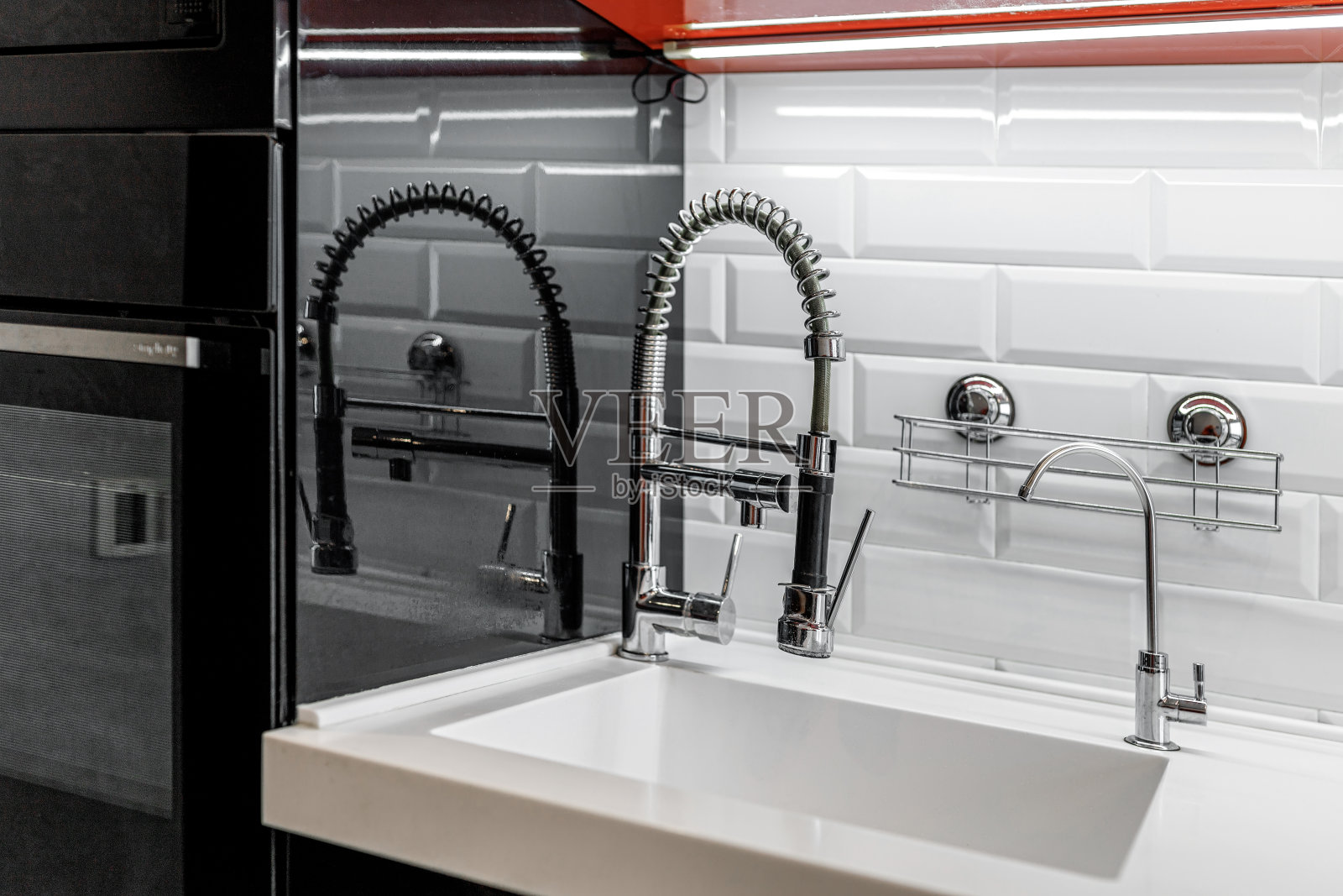 厨房的水龙头是黑色的，靠近墙壁，有光滑的白色砖块和光滑的黑色和红色橱柜照片摄影图片