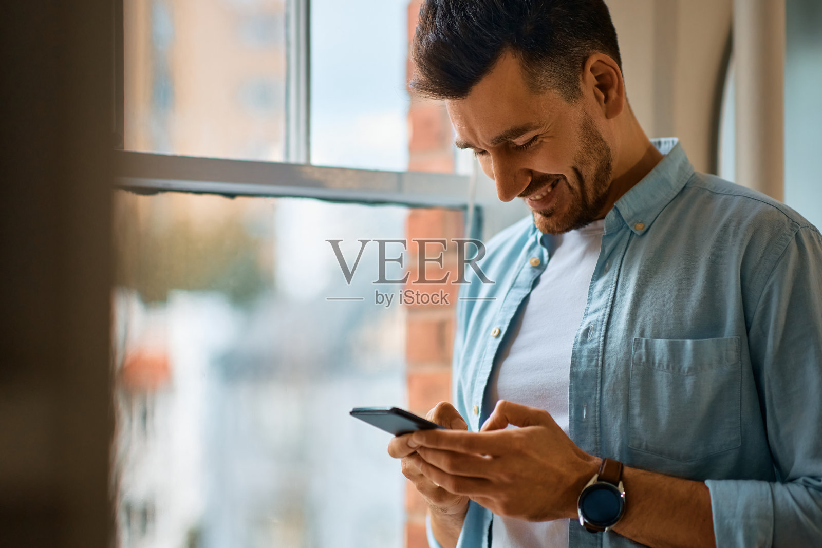快乐男人在窗边用智能手机发短信。照片摄影图片