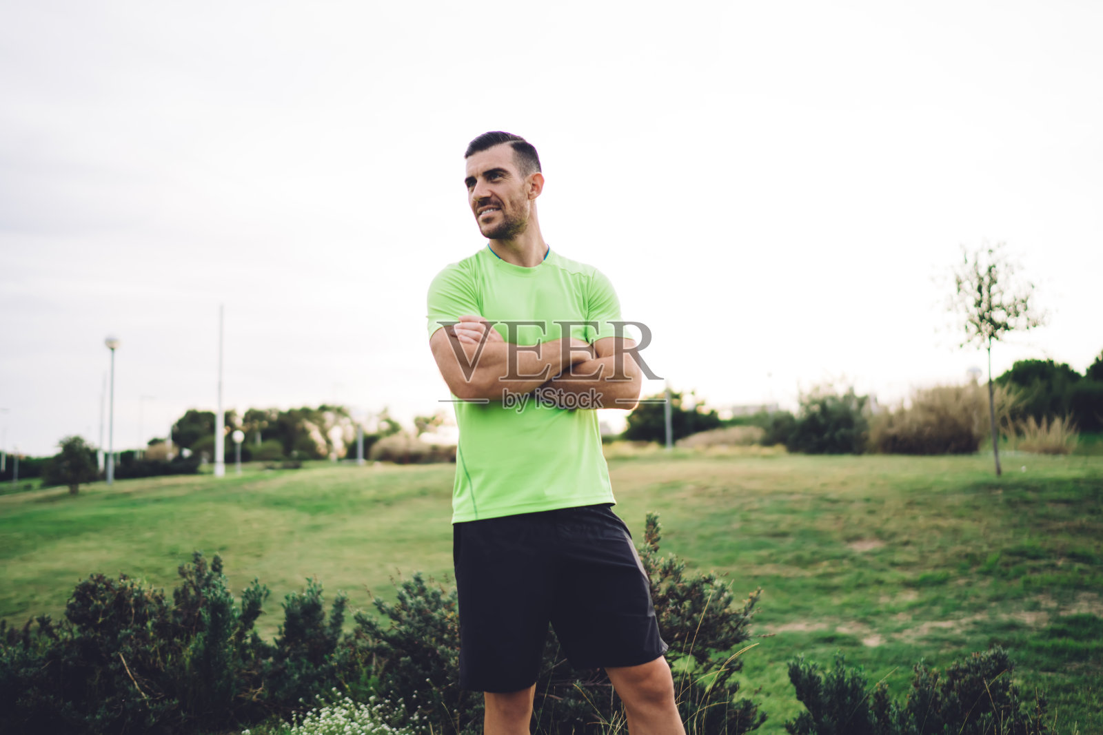 快乐的白人男性教练，交叉肌肉的双手在公园锻炼时微笑，拥有完美身材的成功运动员，保持健康的生活方式和阅读运动目标照片摄影图片