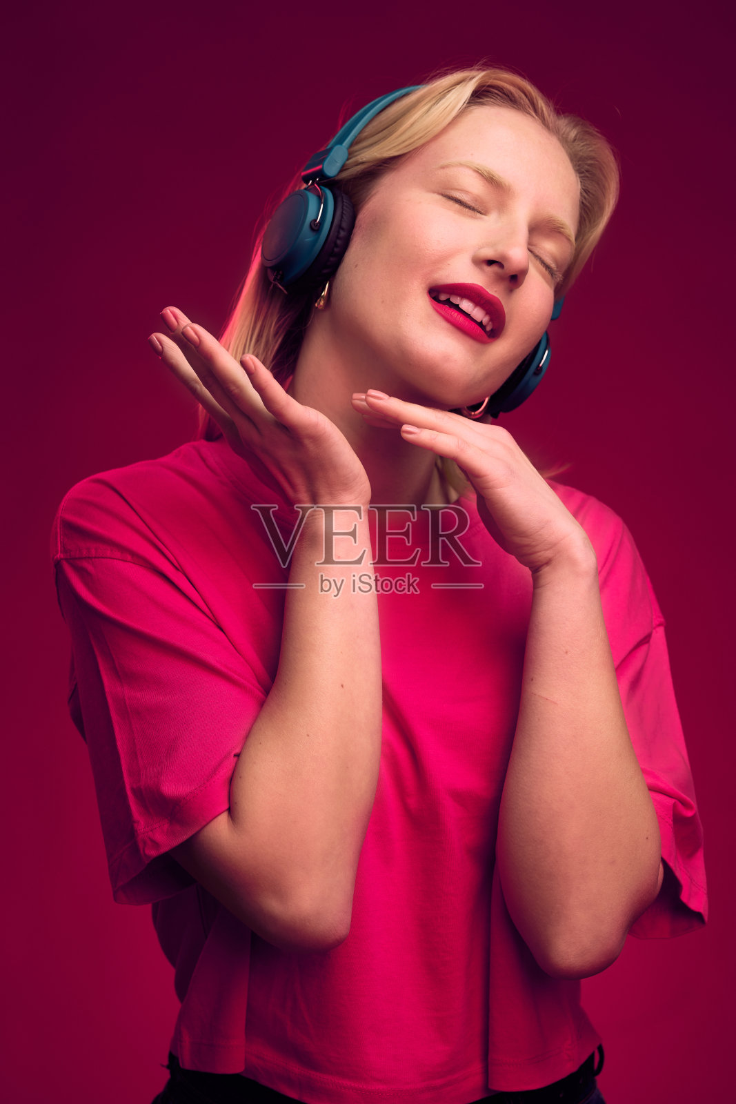 一个美丽的金发女人享受轻松的音乐耳机，站在洋红色的背景孤立。洋红色万岁，年度流行色。照片摄影图片