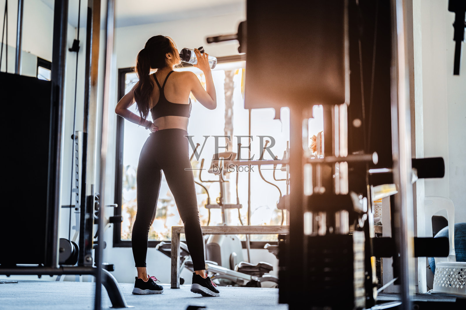 健身亚洲女子在锻炼后放松她在健身房喝水。美丽的年轻女子站在健身房看向别处。健身，健美，健康的生活方式概念。照片摄影图片