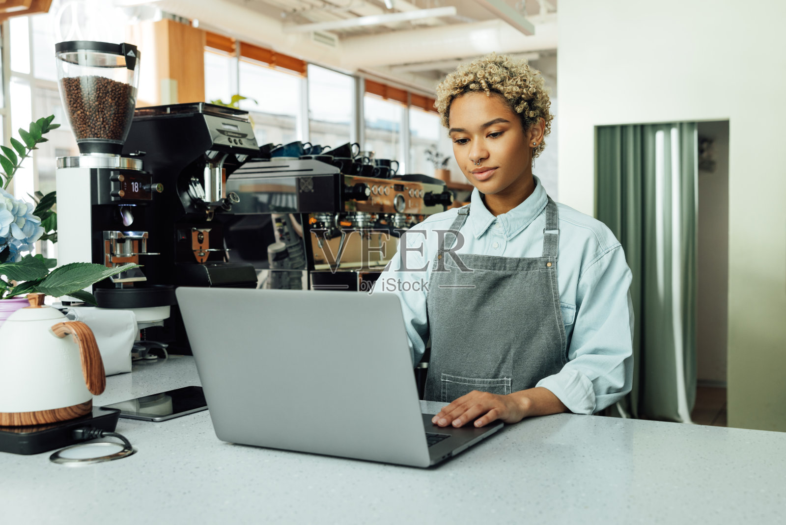 年轻的女咖啡师在咖啡店用笔记本电脑工作。一个围着围裙的短发女人在她当地的小咖啡馆里。照片摄影图片