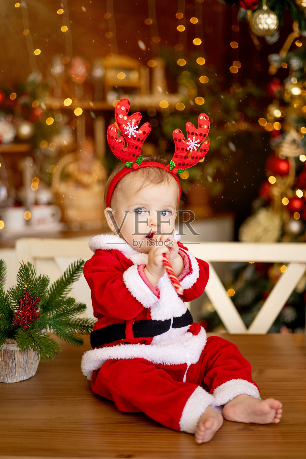 一个小孩穿着圣诞老人的服装，戴着有趣的鹿耳朵，坐在厨房里的节日桌子上，桌子上有圣诞树和礼物照片摄影图片