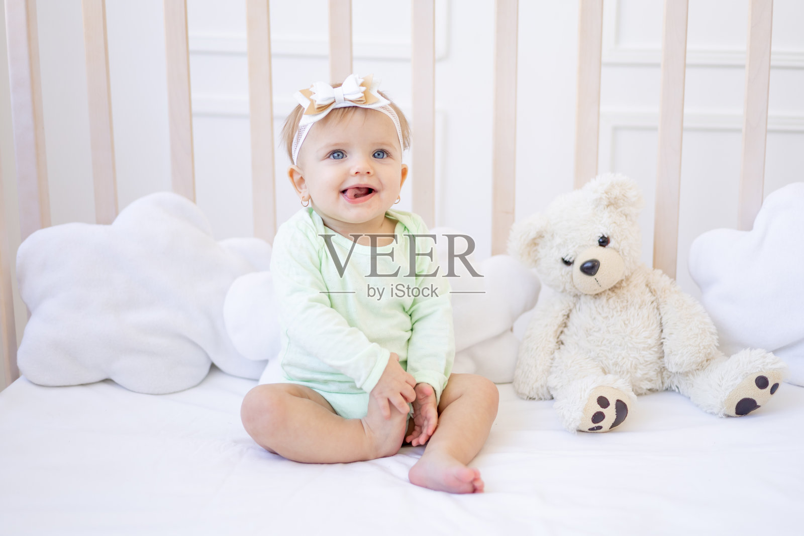 可爱有趣的小女婴在儿童明亮的房间里，穿着绿色紧身衣，在白色棉床上的婴儿床上露出舌头，微笑着照片摄影图片