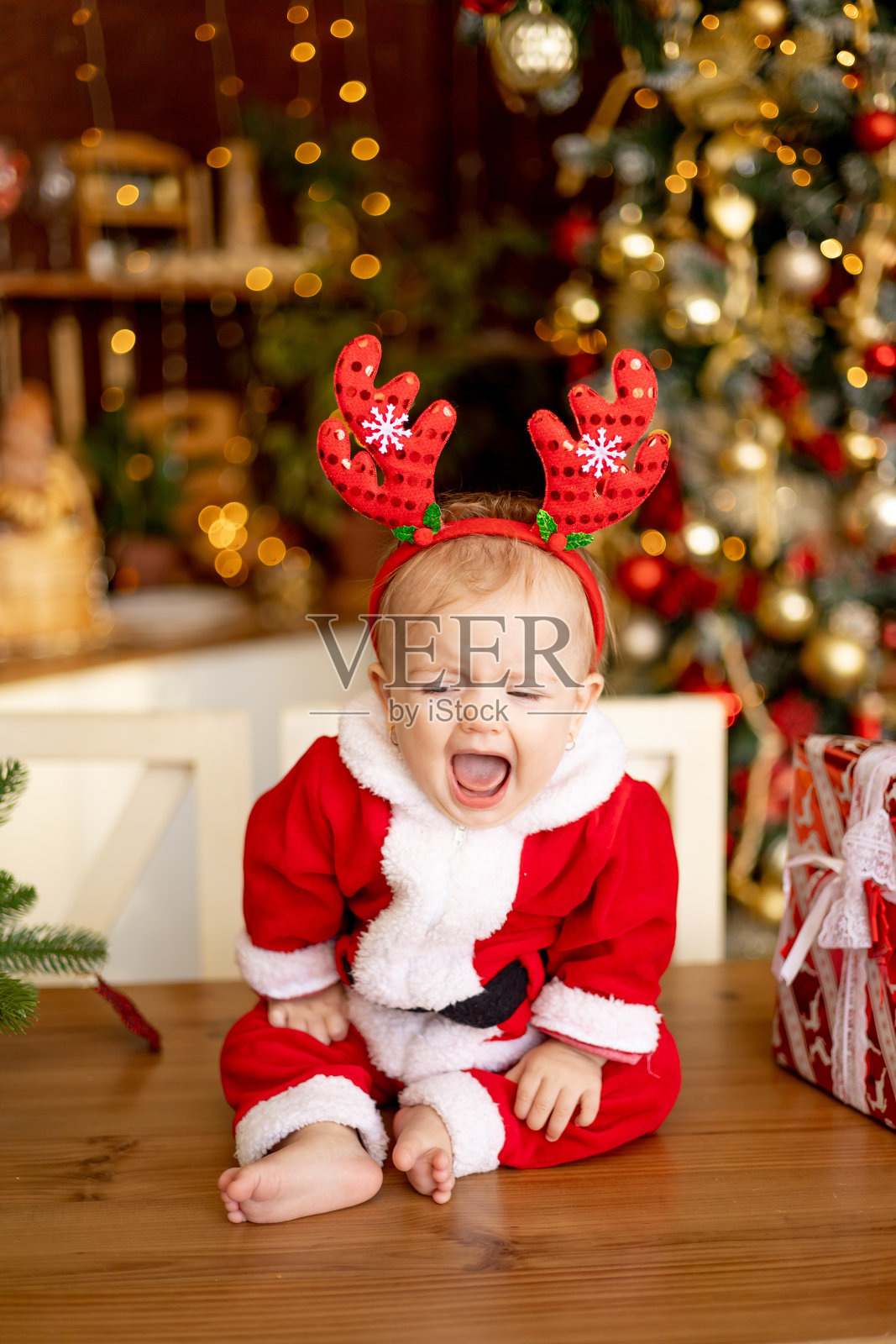 一个小孩穿着圣诞老人的服装，戴着有趣的鹿耳朵，坐在厨房里的节日桌子上，桌子上有圣诞树和礼物照片摄影图片