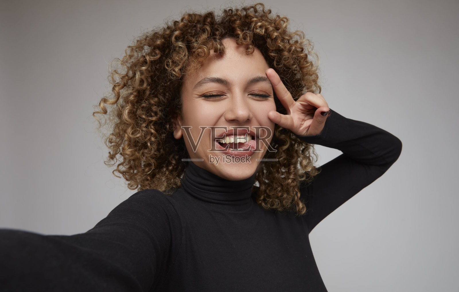 年轻的卷发女子自拍，伸出舌头，做和平手势。照片摄影图片