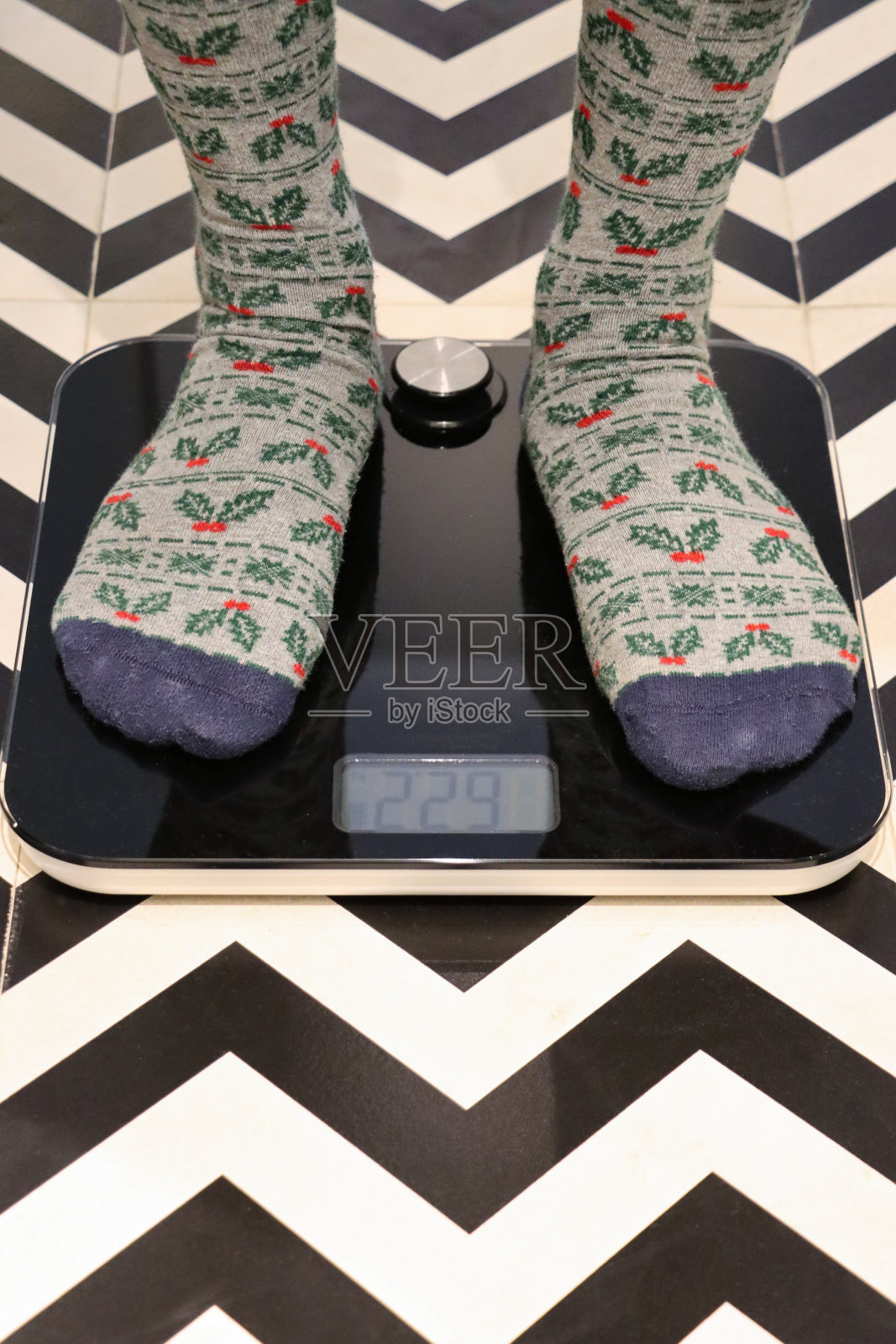 无法辨认的人穿着带有冬青叶和浆果图案的圣诞图案袜子站在体重秤上，豪华酒店浴室单色锯齿形图案地砖照片摄影图片
