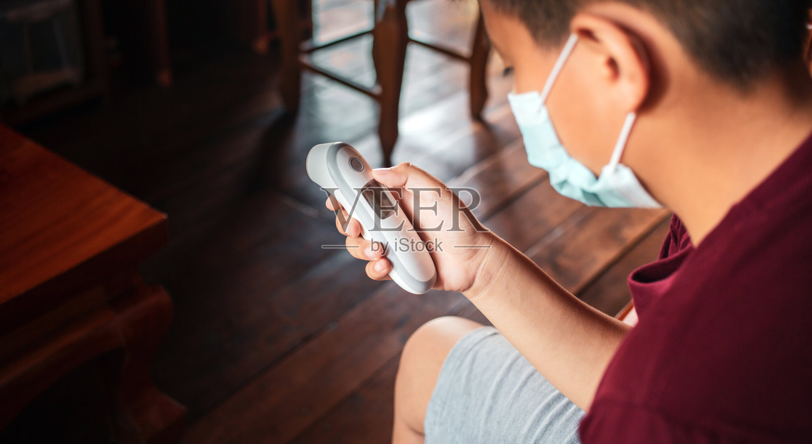 戴着口罩的亚洲男孩在家用红外线体温计测体温，看起来很不舒服照片摄影图片