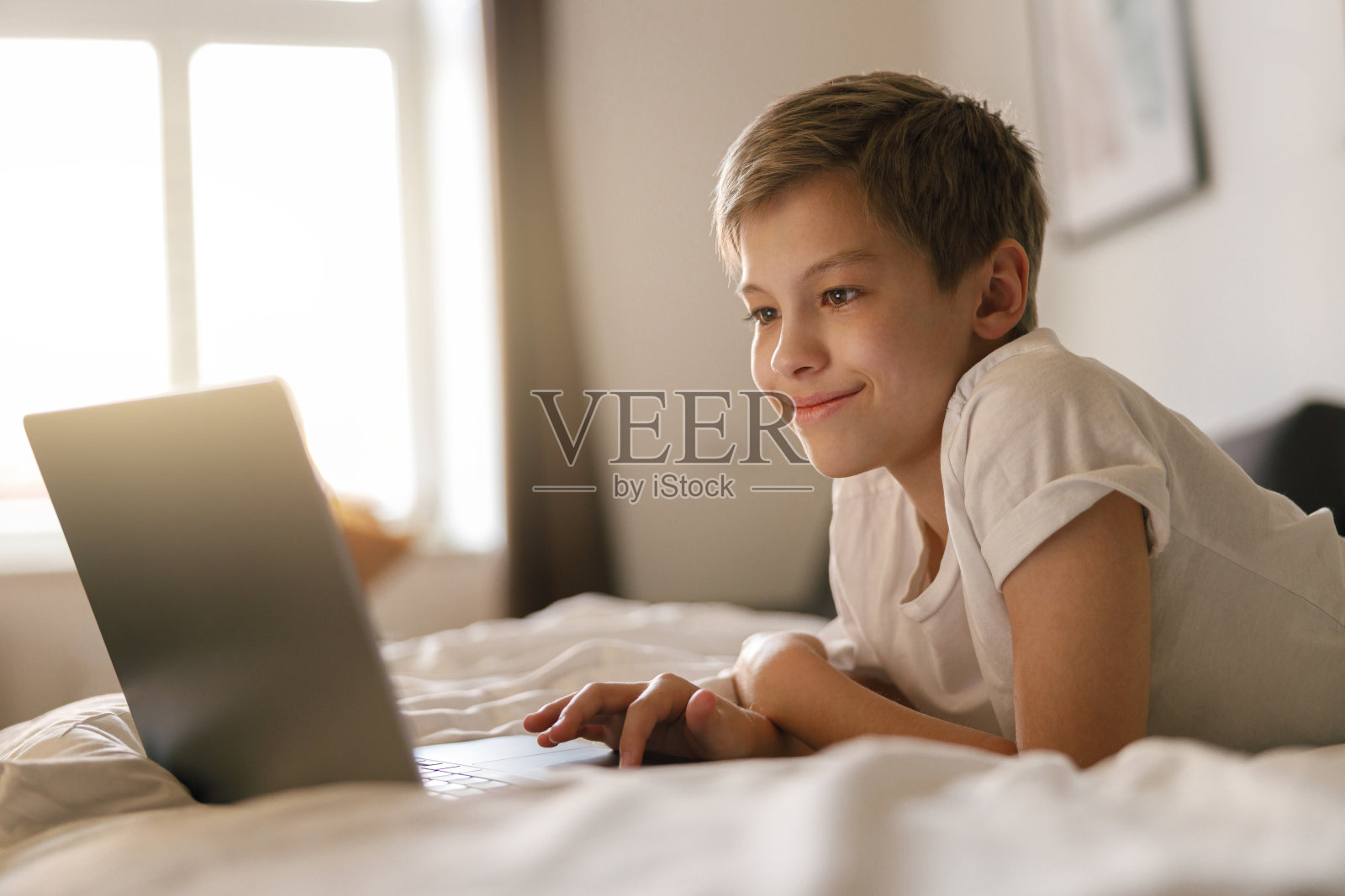 小男孩躺在床上，看着打开的笔记本电脑。家庭教育理念照片摄影图片
