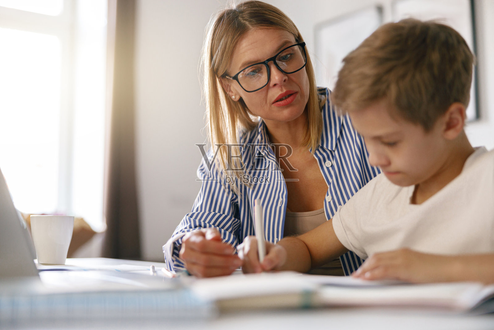 男孩坐在桌子上写在网上课程的抄写本，而他的妈妈帮助他照片摄影图片