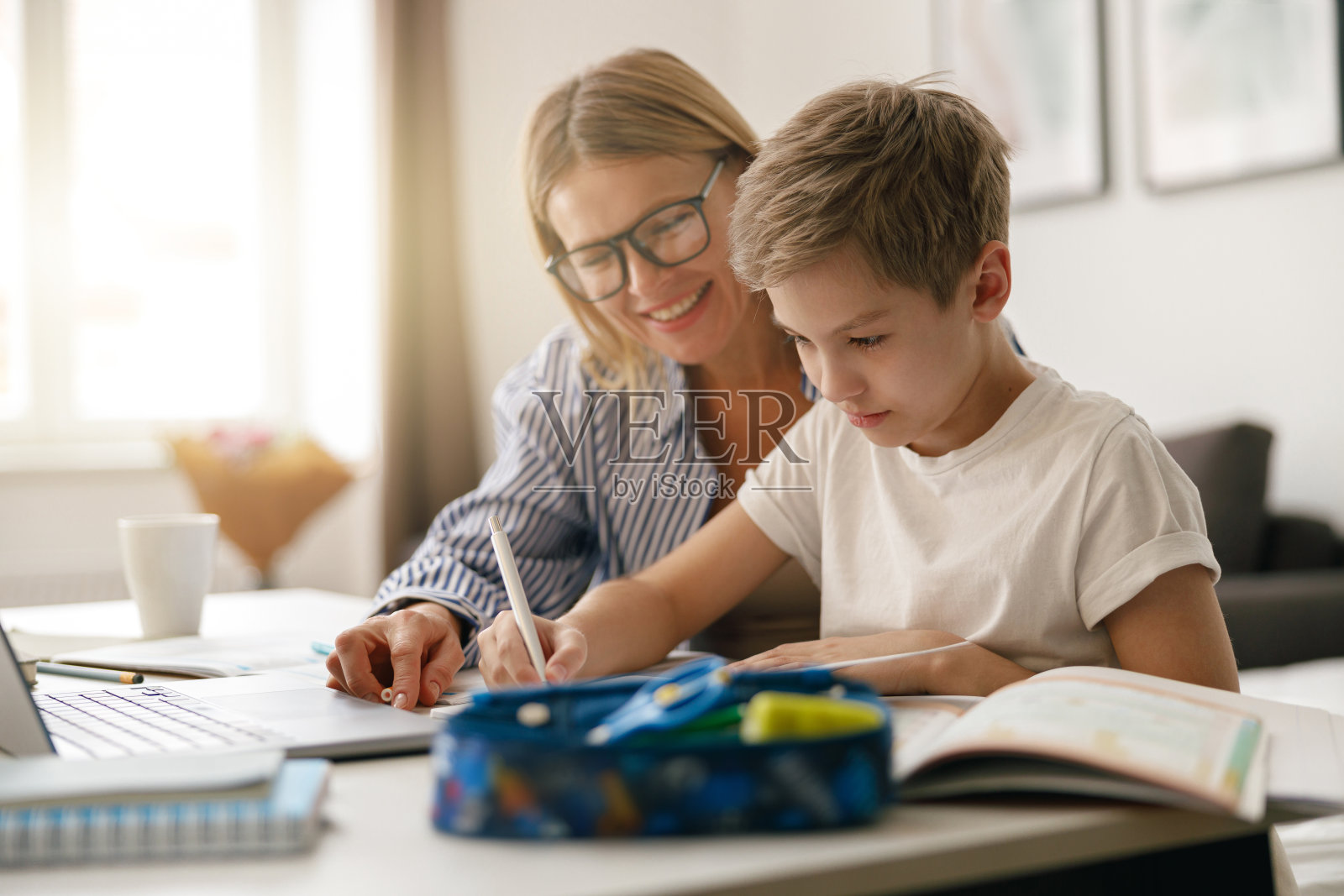 男孩坐在桌子上写在网上课程的抄写本，而他的妈妈帮助他照片摄影图片