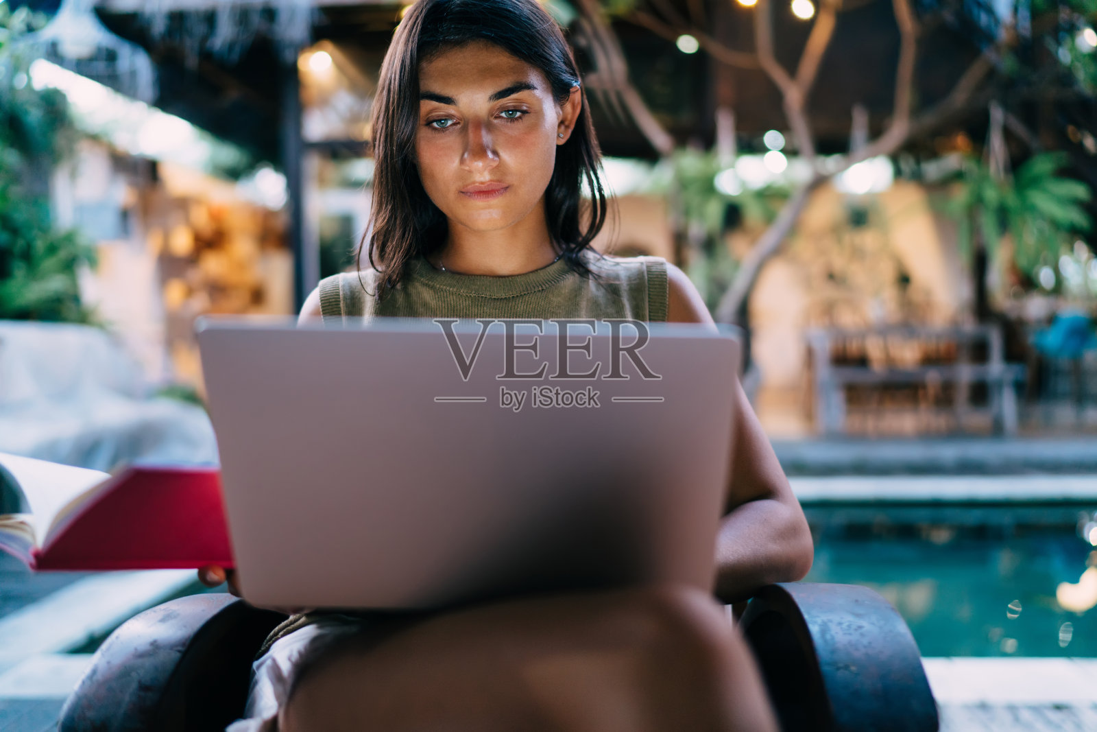 严肃的年轻民族女性自由职业者在笔记本电脑上打字坐在泳池边的阳台照片摄影图片