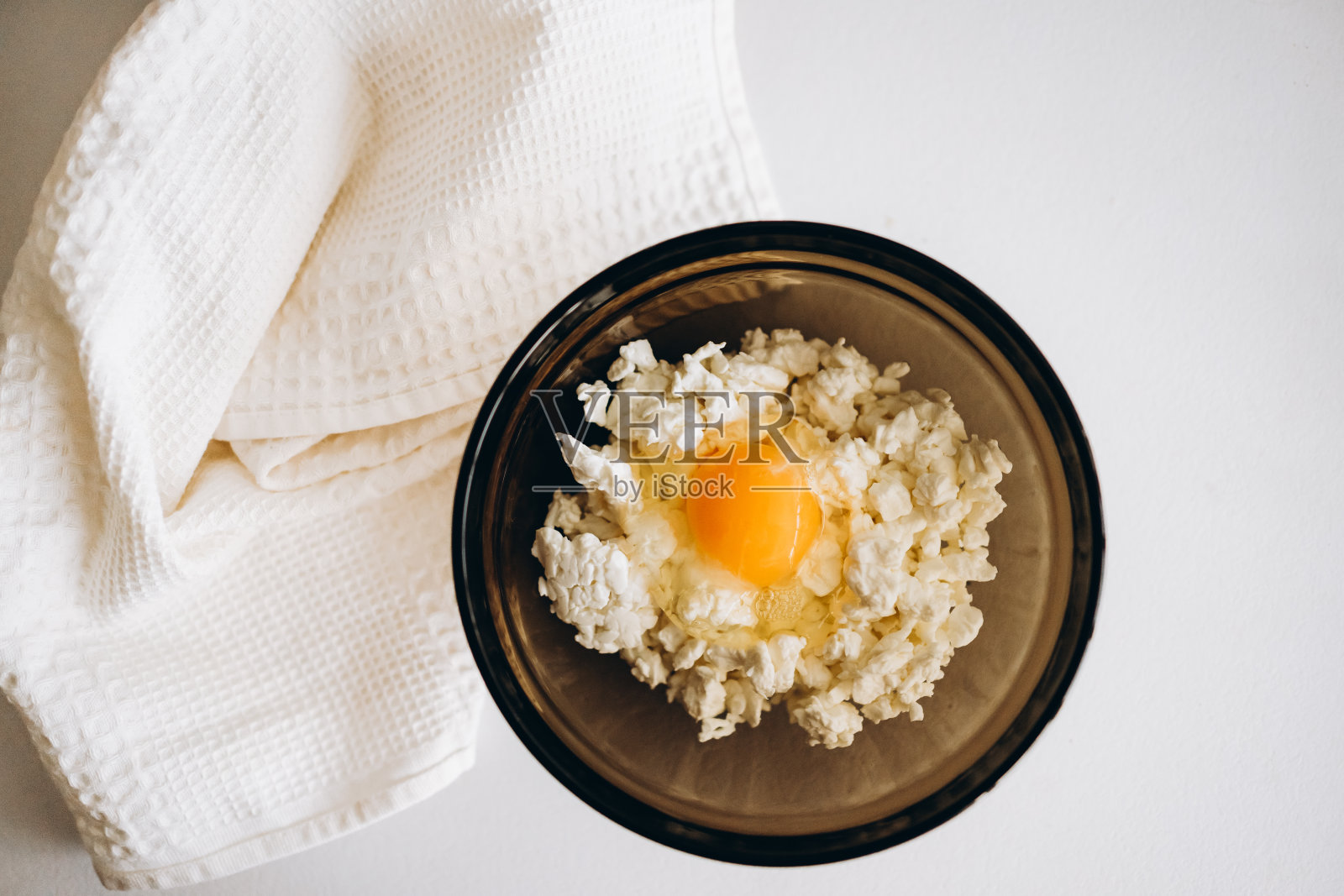 白桌子上的黑色玻璃碗里装着白干酪、鸡蛋和盐。轻毛巾放在桌上。亲手做早餐照片摄影图片