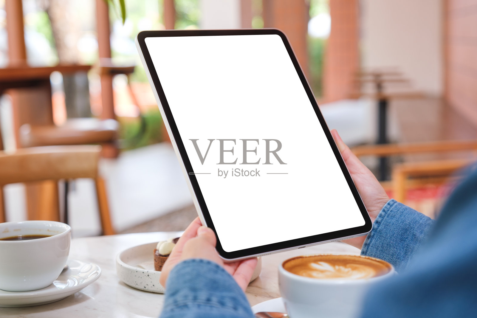 在咖啡馆的桌子上，一个女人拿着空白白色桌面屏幕的数字平板电脑和蛋糕和咖啡杯的模型图像照片摄影图片