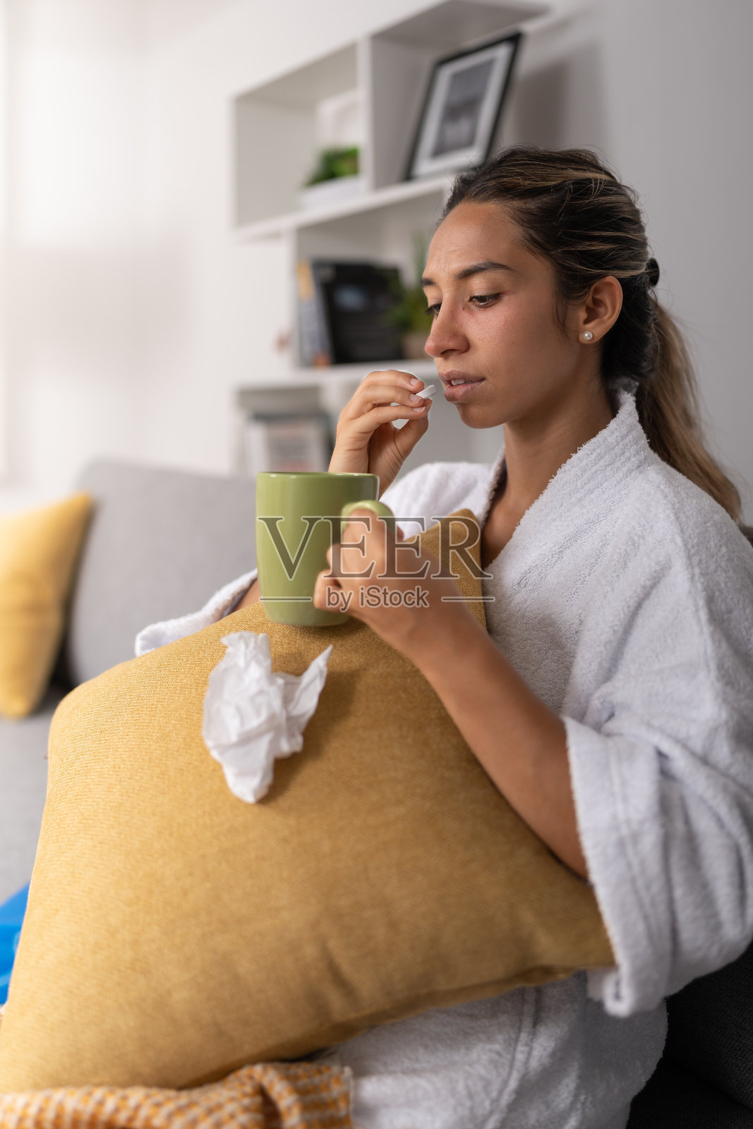 生病的年轻女子，一边吃着药丸，一边端着一杯绿茶，坐在客厅里照片摄影图片
