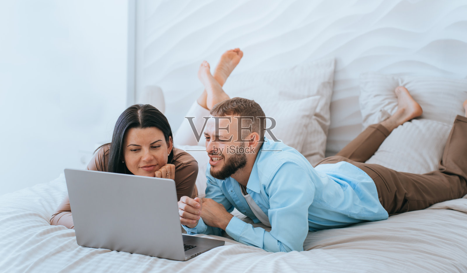 年轻的白人夫妇在休闲躺在床上用笔记本电脑通过互联网观看视频。愉快的西班牙女孩放松与男友在周末回家。国内休闲。新婚夫妇拥有新电脑。照片摄影图片