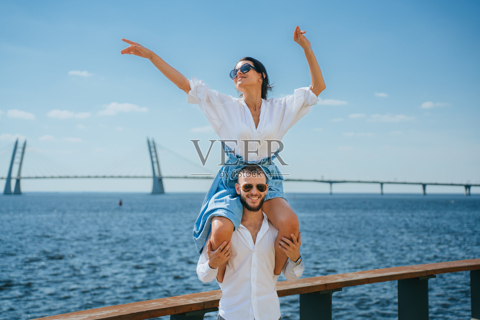 快乐的西班牙年轻女子在太阳镜和牛仔裤裙子坐在男朋友的肩膀上挥手对海洋和桥。白种夫妇在海边旅行，在度假胜地度蜜月，旅行。照片摄影图片